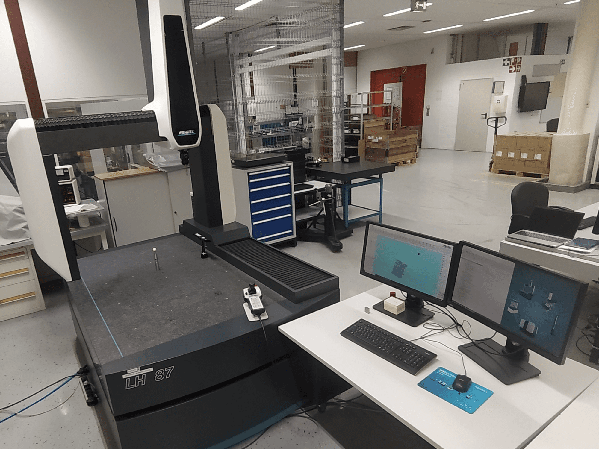 Máquina de medição por coordenadas Wenzel LH 87 visível num laboratório, com dois monitores e sonda de medição sobre a mesa.