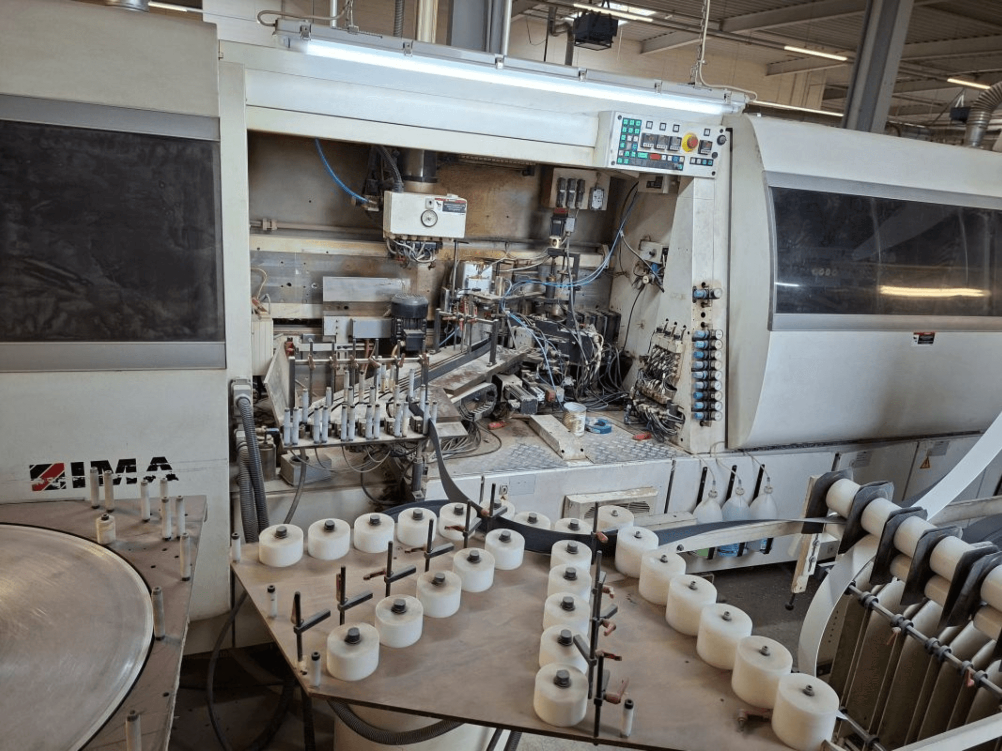 As máquinas IMA, vistas de lado, mostram componentes mecânicos complexos e uma mesa redonda com carretéis brancos.