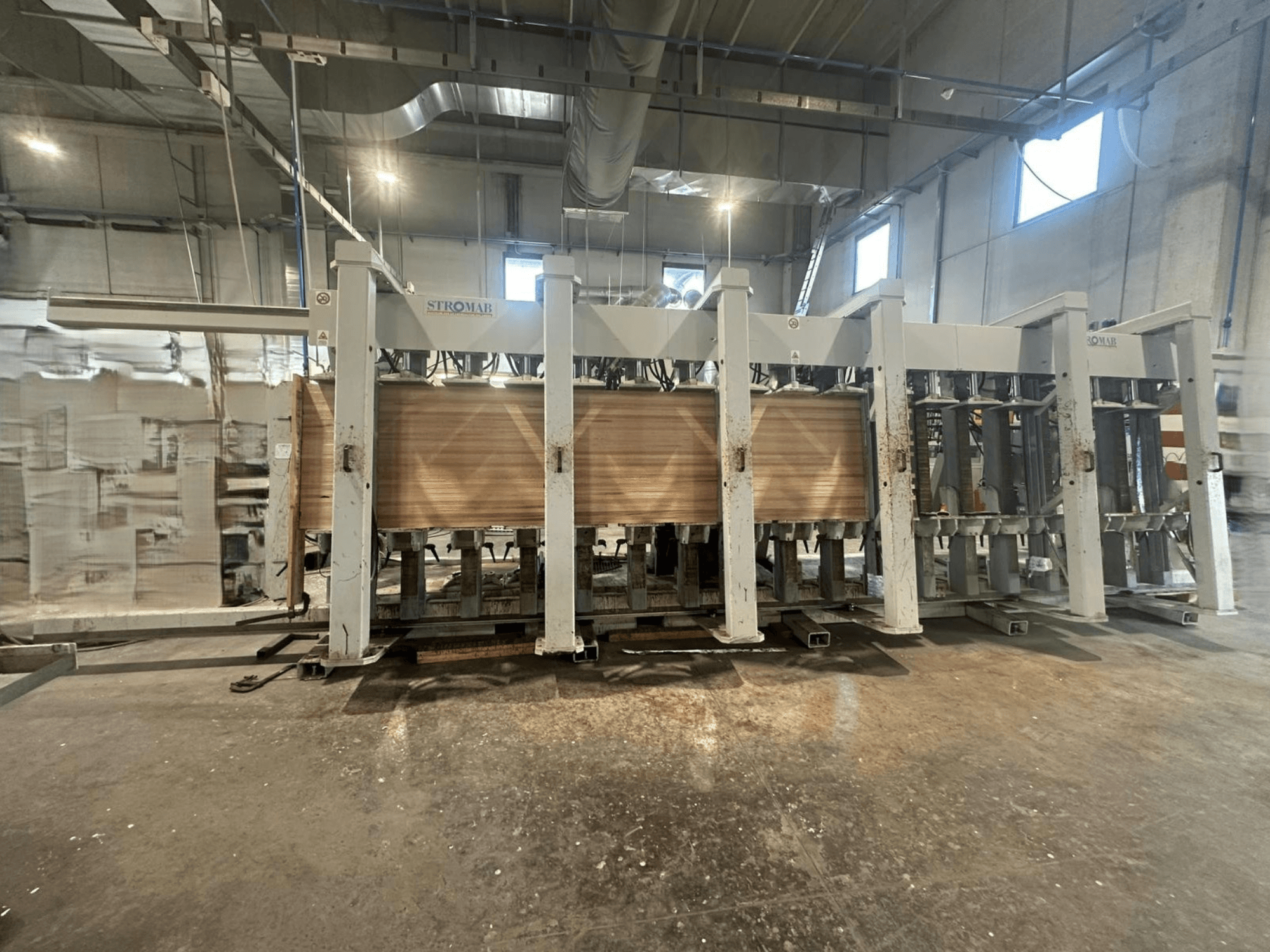 Máquina industrial STROMAB num armazém, vista frontal, com painéis de madeira a serem processados, mostrando a estrutura metálica e as peças mecânicas.