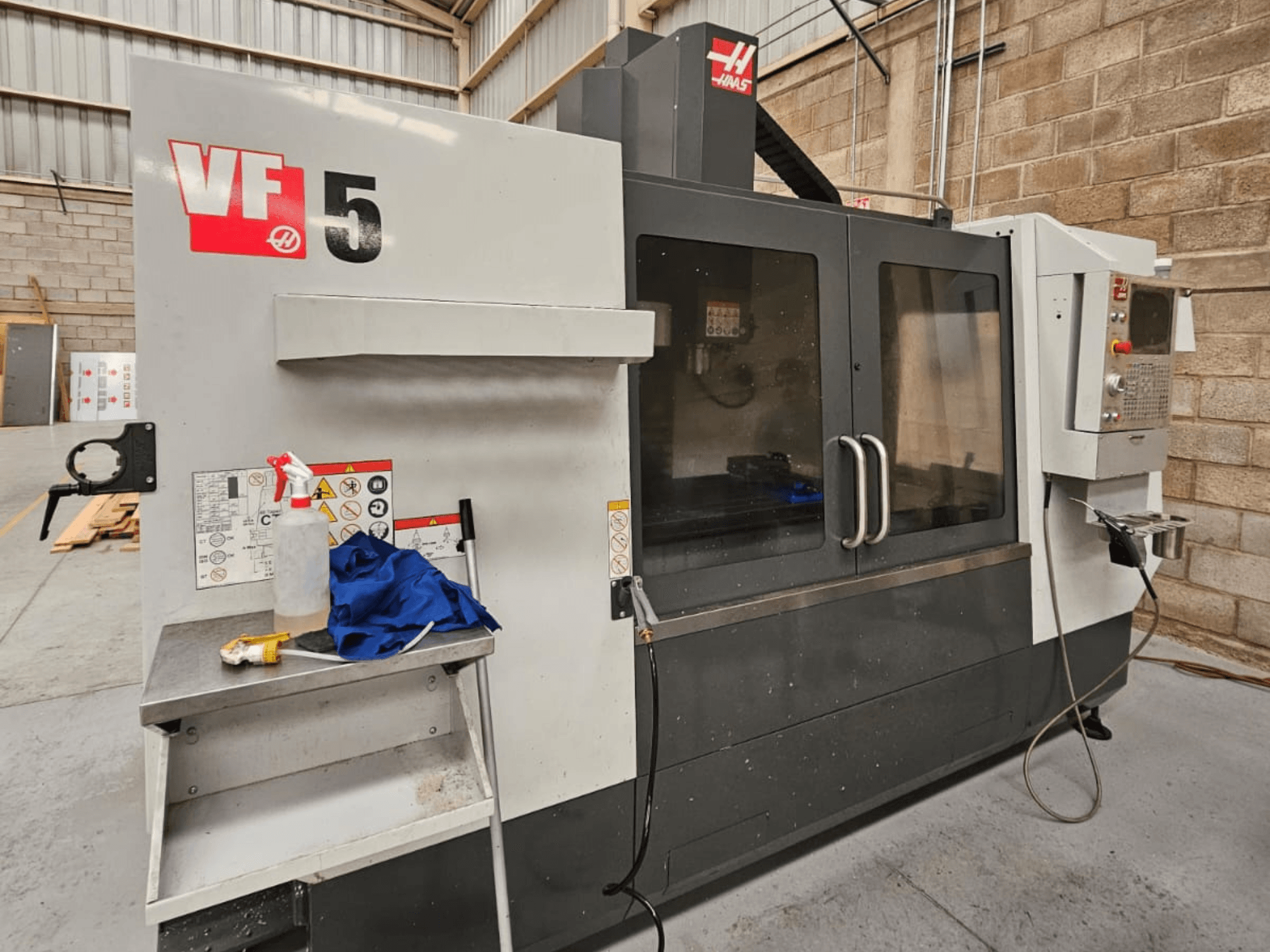 Fresadora CNC Haas VF-5 em vista frontal, com painel de controlo, garrafa de líquido de refrigeração, pano azul e instruções de segurança.
