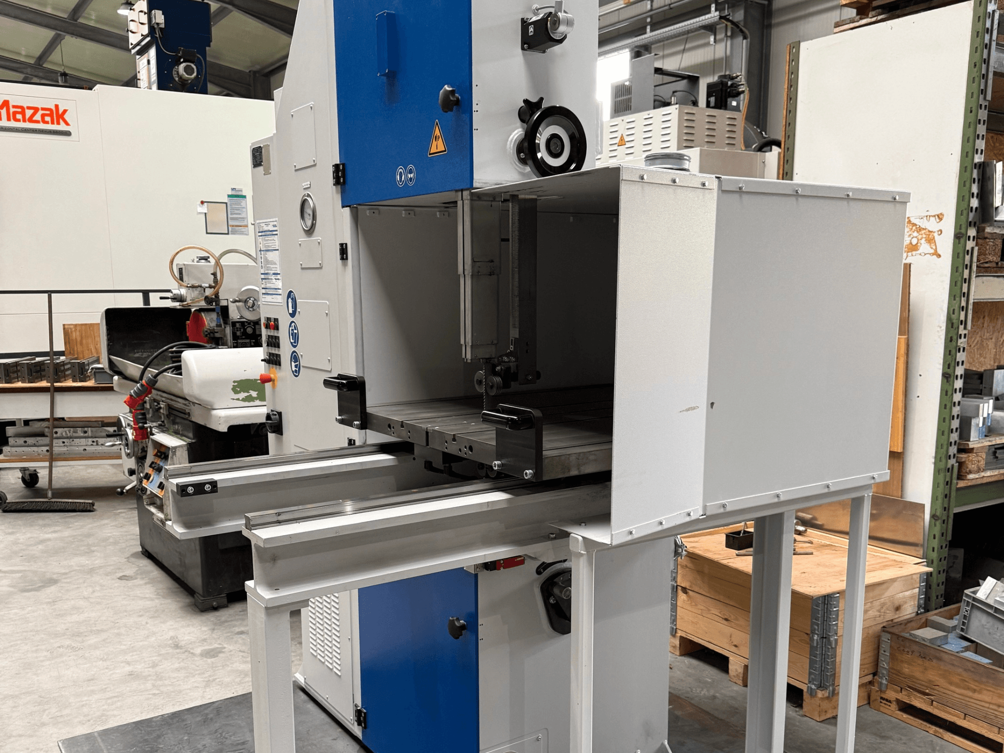 Máquina industrial Mazak vista de frente, com um painel de controlo azul, mesa metálica e avisos de segurança.