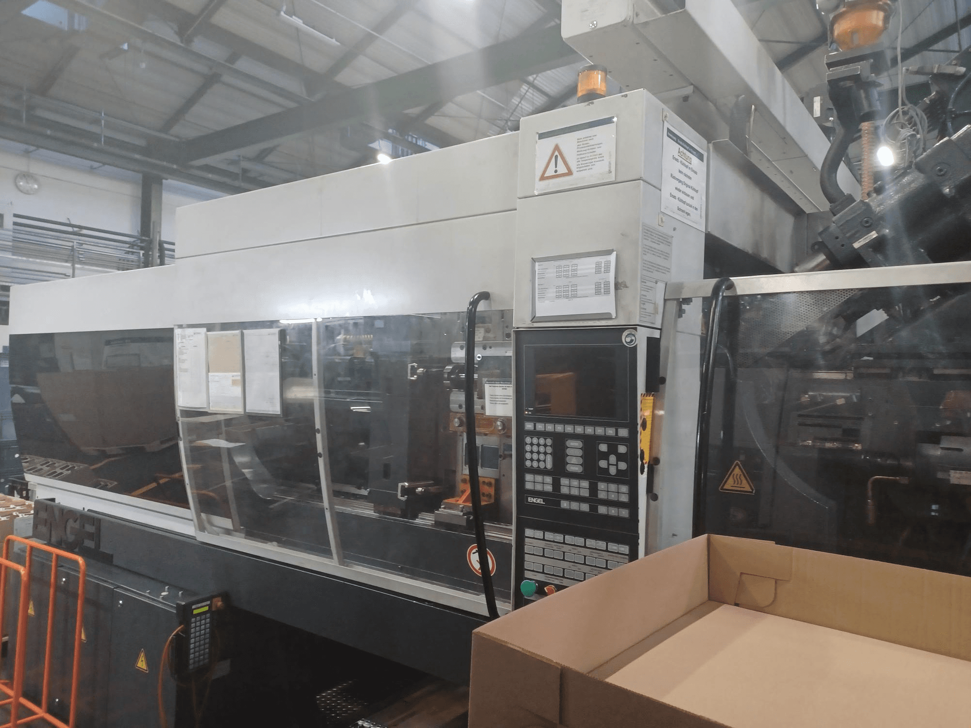 Máquina de moldagem por injeção Engel, vista frontal, com painel de controlo, etiquetas de segurança e uma caixa vazia posicionada nas proximidades.