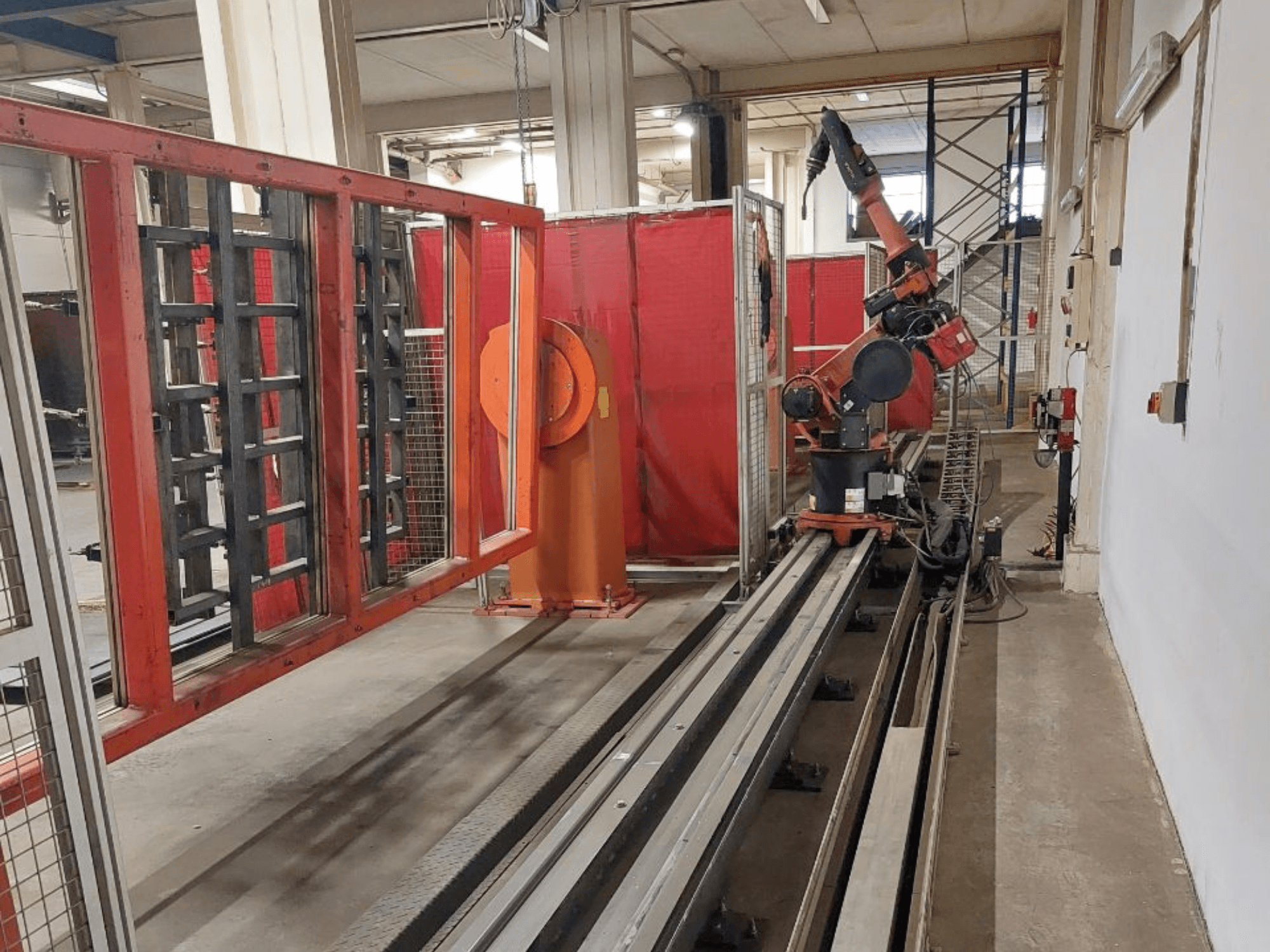 Braço robótico industrial com cores laranja e preto posicionado em calhas metálicas, rodeado por barreiras de proteção vermelhas, vista frontal.