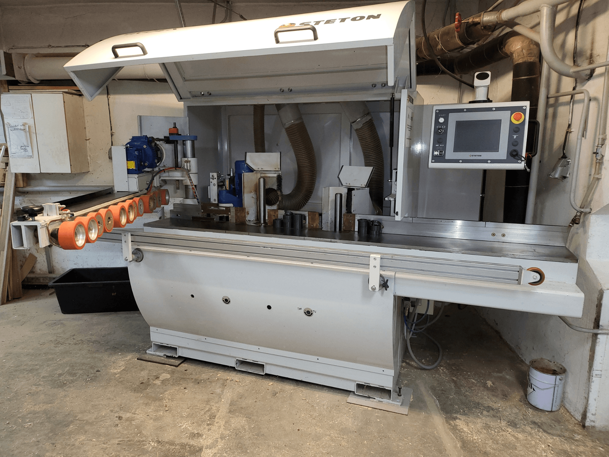 Vista frontal de uma máquina de trabalhar madeira Stetton com um painel de controlo, tubos de alimentação e várias peças de madeira sobre a mesa.