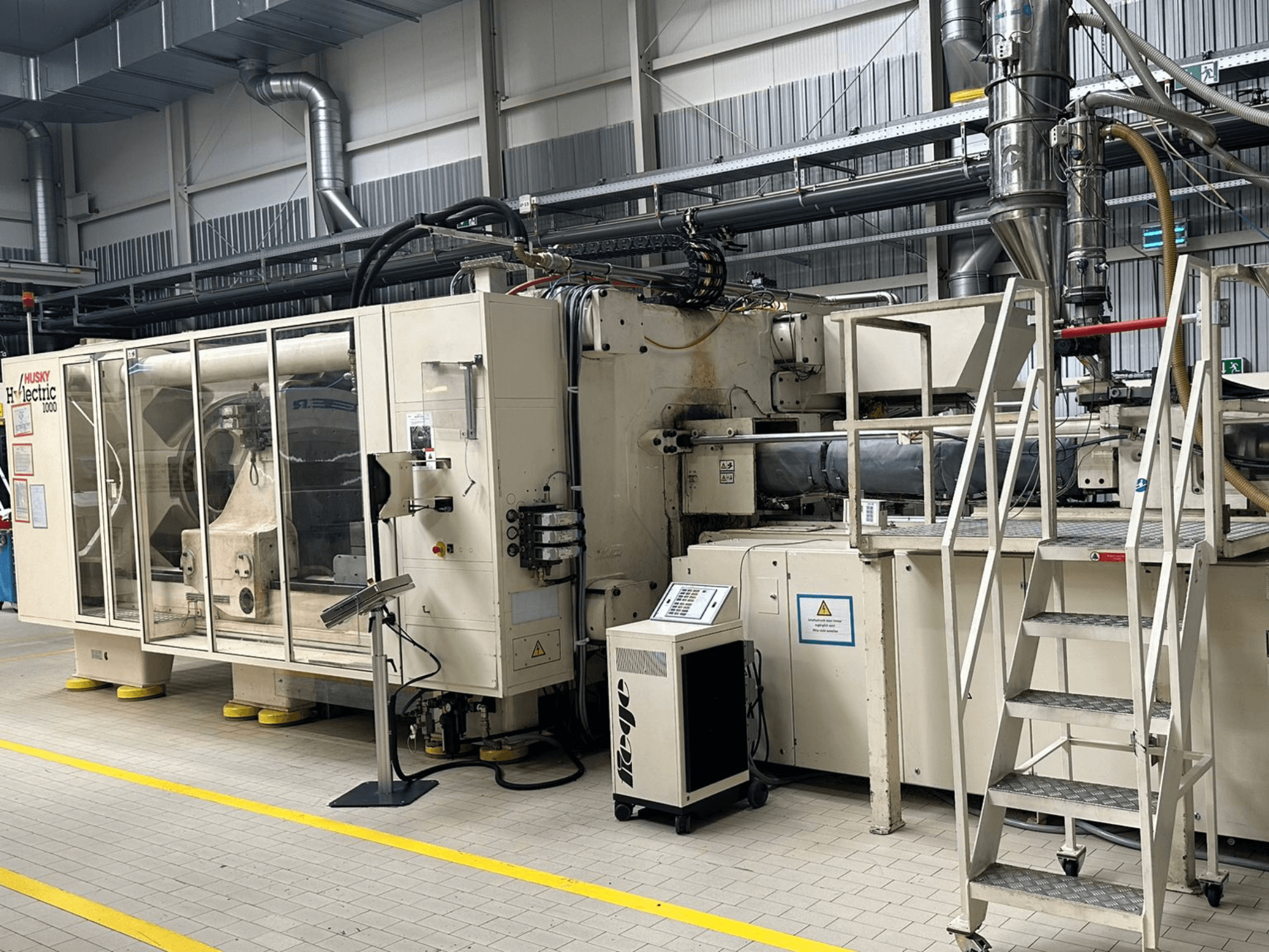 Vista frontal da máquina de moldagem por injeção elétrica Husky Hylelectric H1000 RS95/80 num ambiente fabril, apresentando o painel de controlo e os degraus.