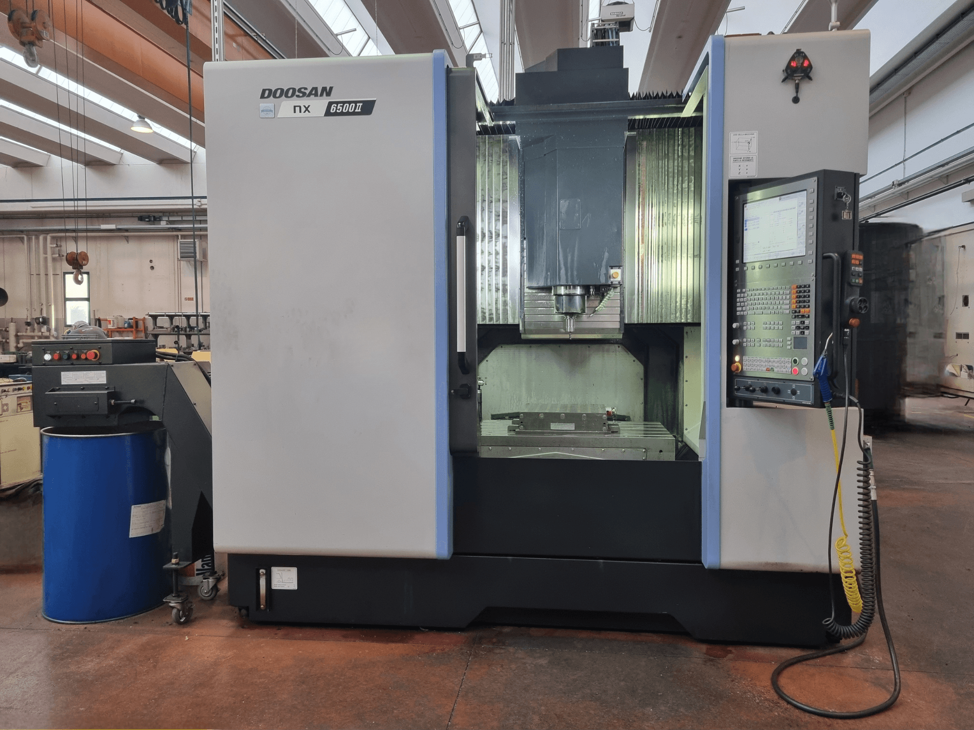 Máquina CNC Doosan NX 6500 II, vista frontal, com painel de controlo e área de maquinagem, posicionada ao lado de um barril azul.