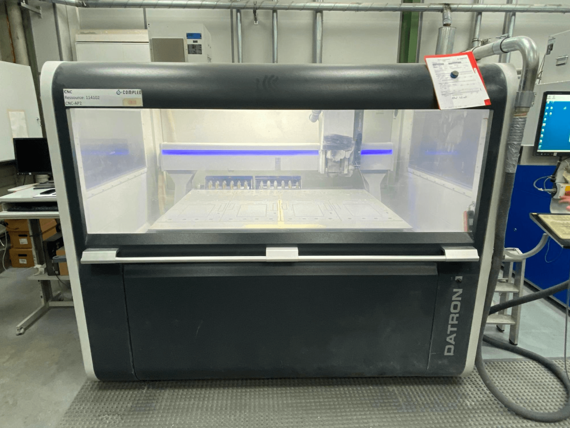 Vista frontal de um centro de maquinação vertical Datron MLCube B, com controlos visíveis e uma mesa de trabalho, num ambiente de oficina.