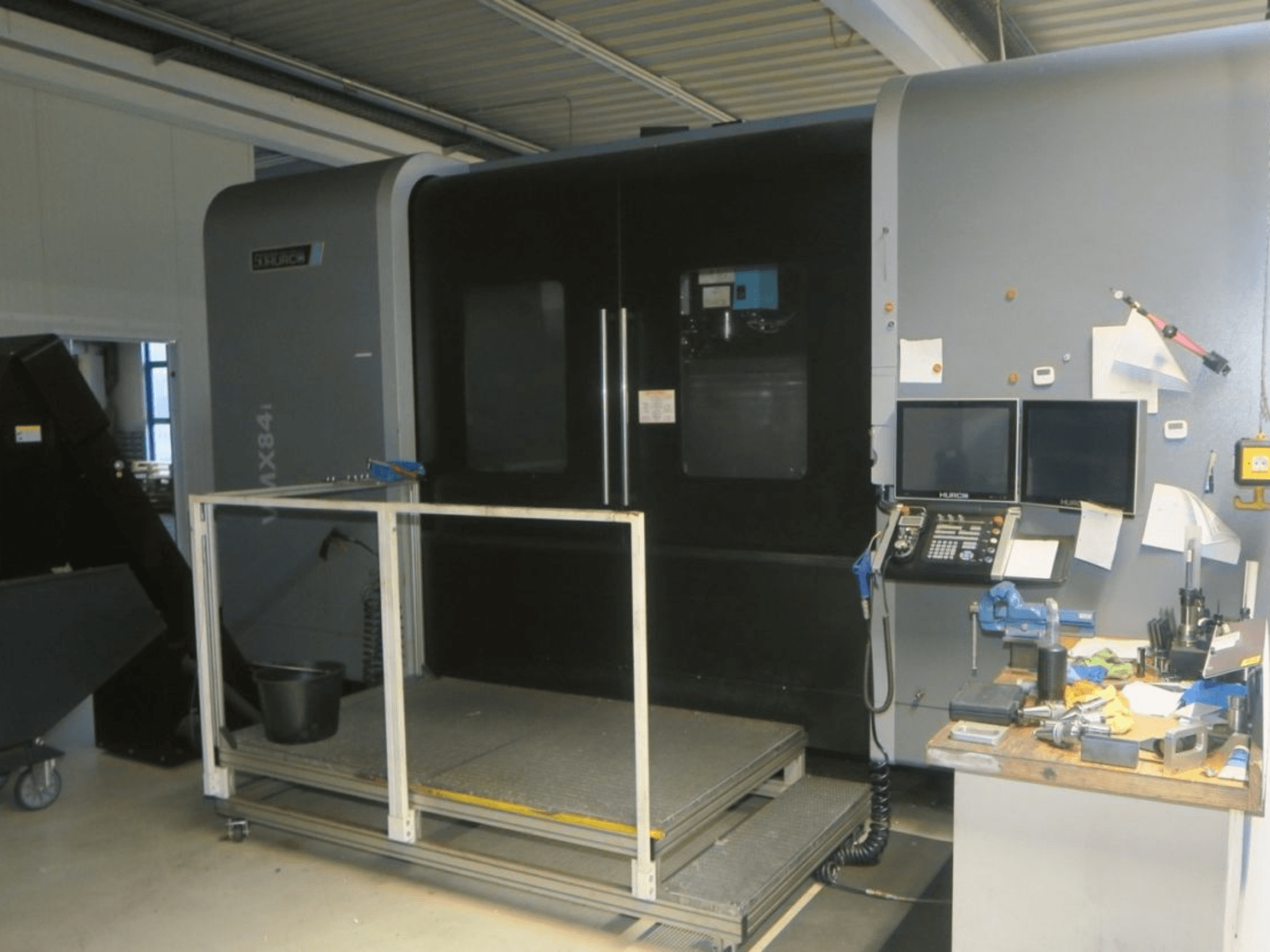 Máquina CNC Hurco MX84 em vista frontal, mostrando monitores duplos, painel de controlo e espaço de trabalho com várias ferramentas.