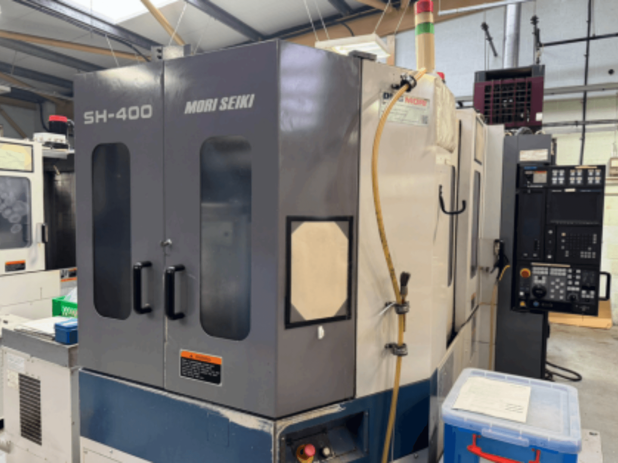 Máquina CNC Mori Seiki SH-400, vista frontal, com portas duplas, painel de controlo e mangueira de fixação. Contentor de armazenamento azul nas proximidades.