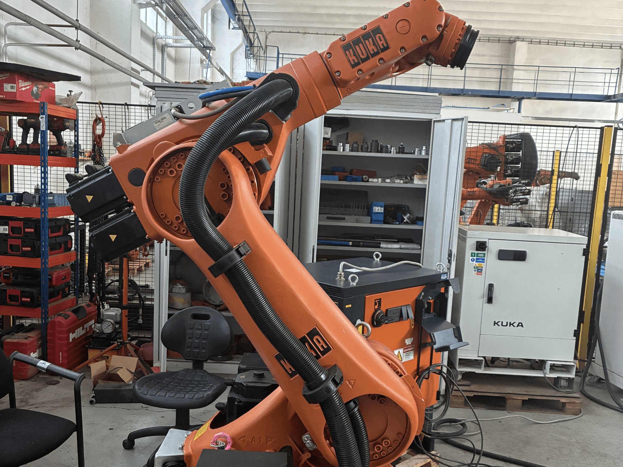 Braço robótico KUKA KR30-3 num ambiente industrial, vista lateral mostrando cabos e controlos, com ferramentas e prateleiras em segundo plano.
