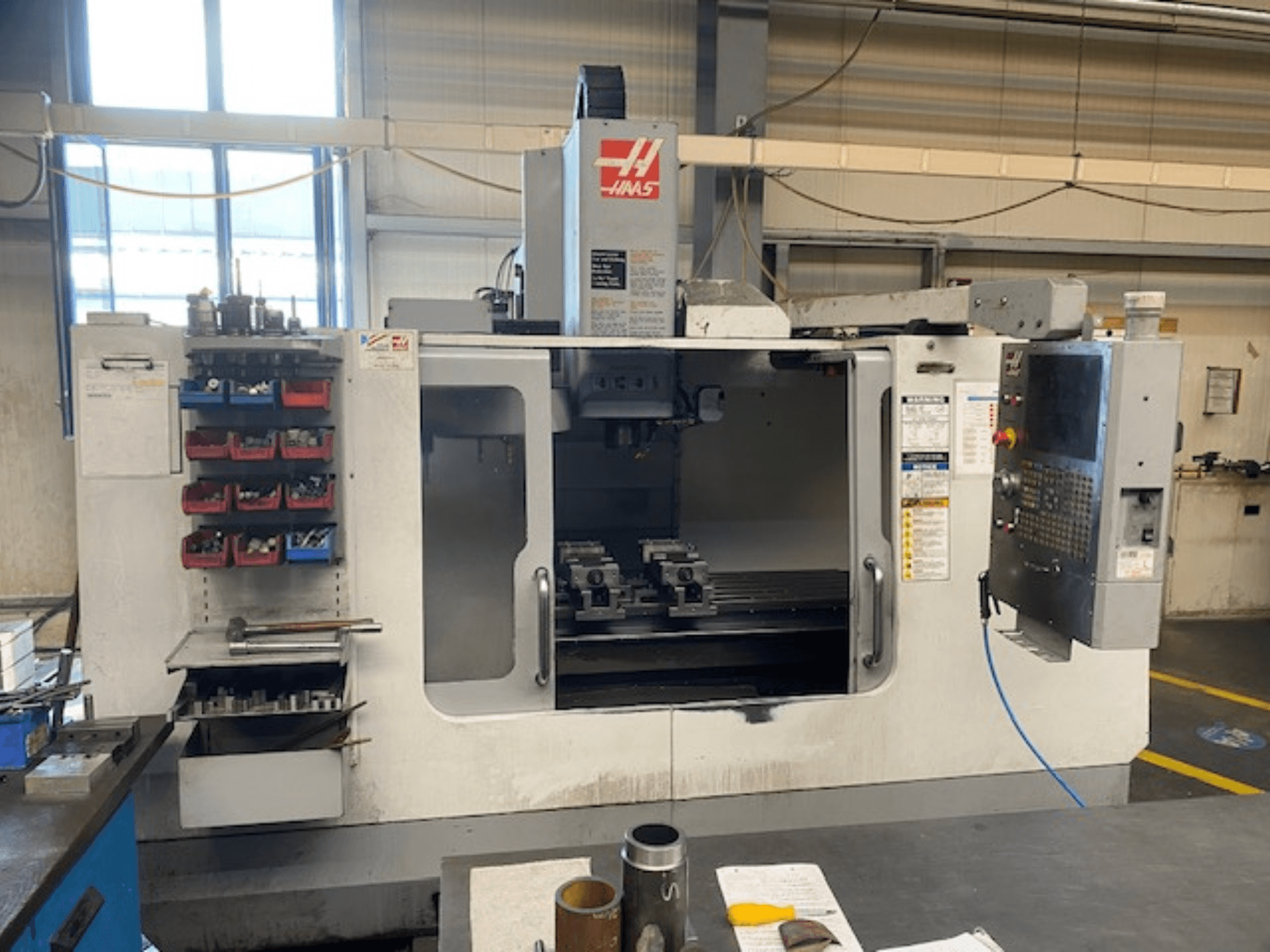 Vista frontal da fresadora CNC Haas, modelo VF-2, com armazenamento de ferramentas, painel de controlo e ferramentas de espaço de trabalho visíveis.