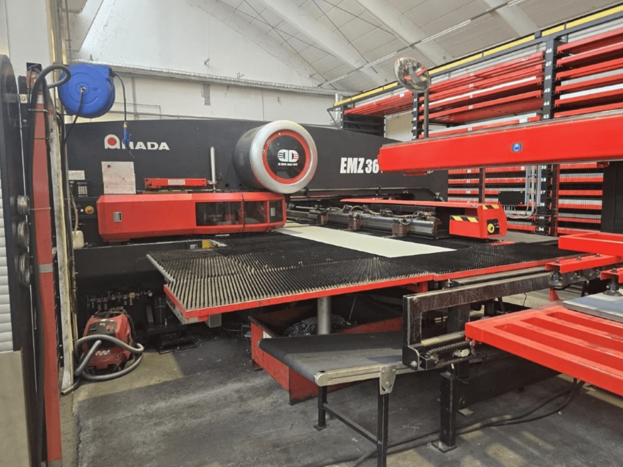 Máquina de corte a laser Amada EMZ 36 numa oficina, vista frontal, com detalhes em vermelho e uma grande mesa de corte.