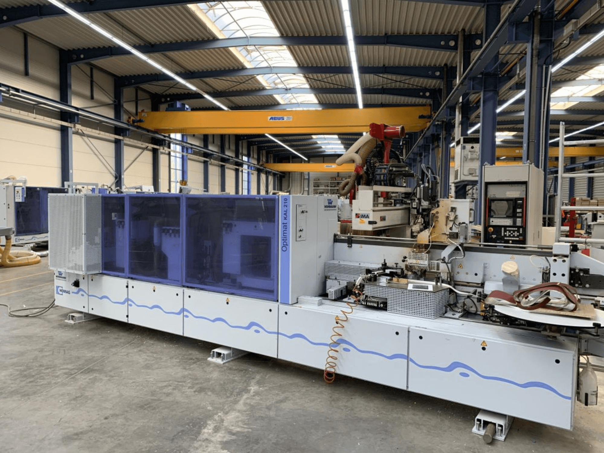 Máquina de encadernação HOMAG Optimat KAL210 vista de frente, mostrando o seu painel de controlo e a área de processamento num ambiente fabril.