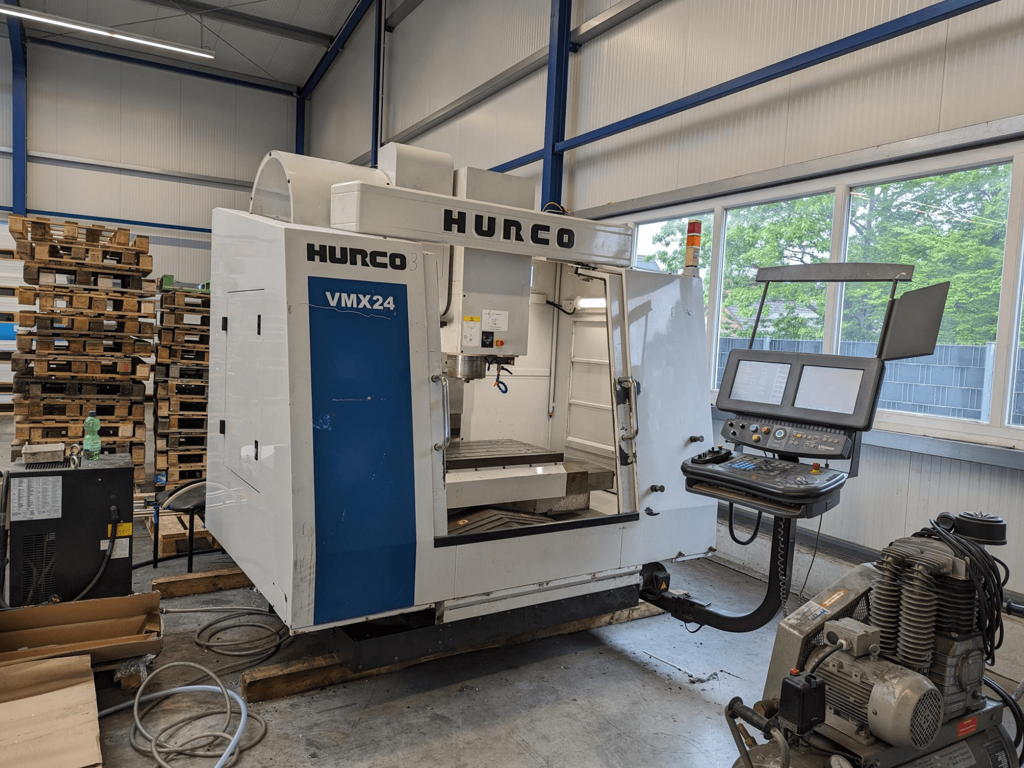 Fresadora Hurco VMX24, vista frontal, com painel de controlo e área de ferramentas; paletes empilhadas em segundo plano; configuração de espaço de trabalho industrial.