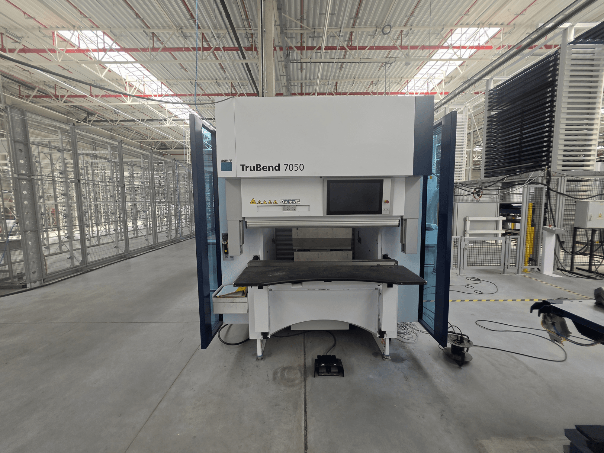Vista frontal da prensa-dobradeira TRUMPF TruBend 7050, mostrando o painel de controlo e a mesa de trabalho num ambiente industrial bem iluminado.