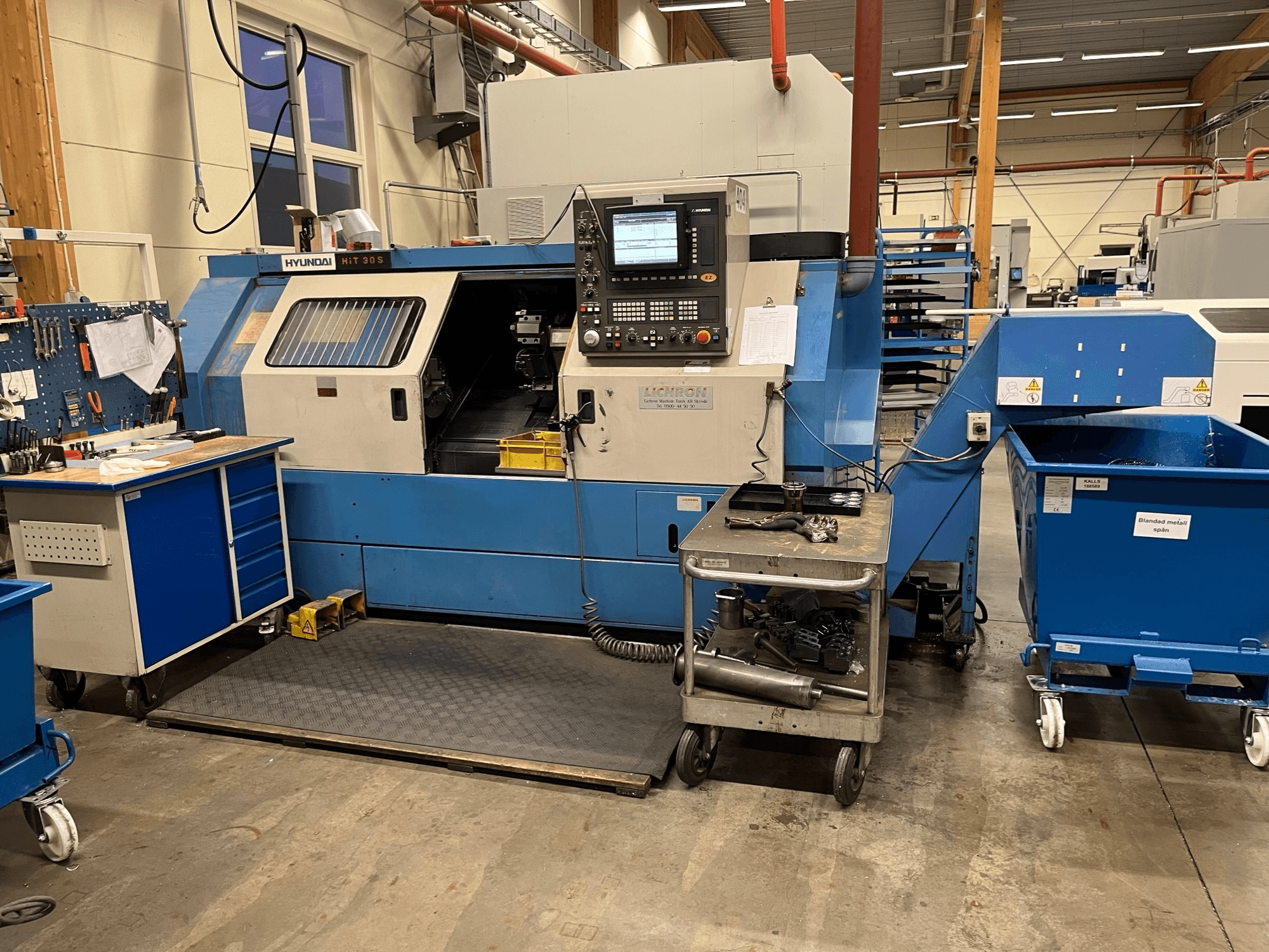 Torneiro horizontal Hyundai HIT 30S, vista frontal, com painel de controlo e ferramentas num carrinho próximo, numa oficina.