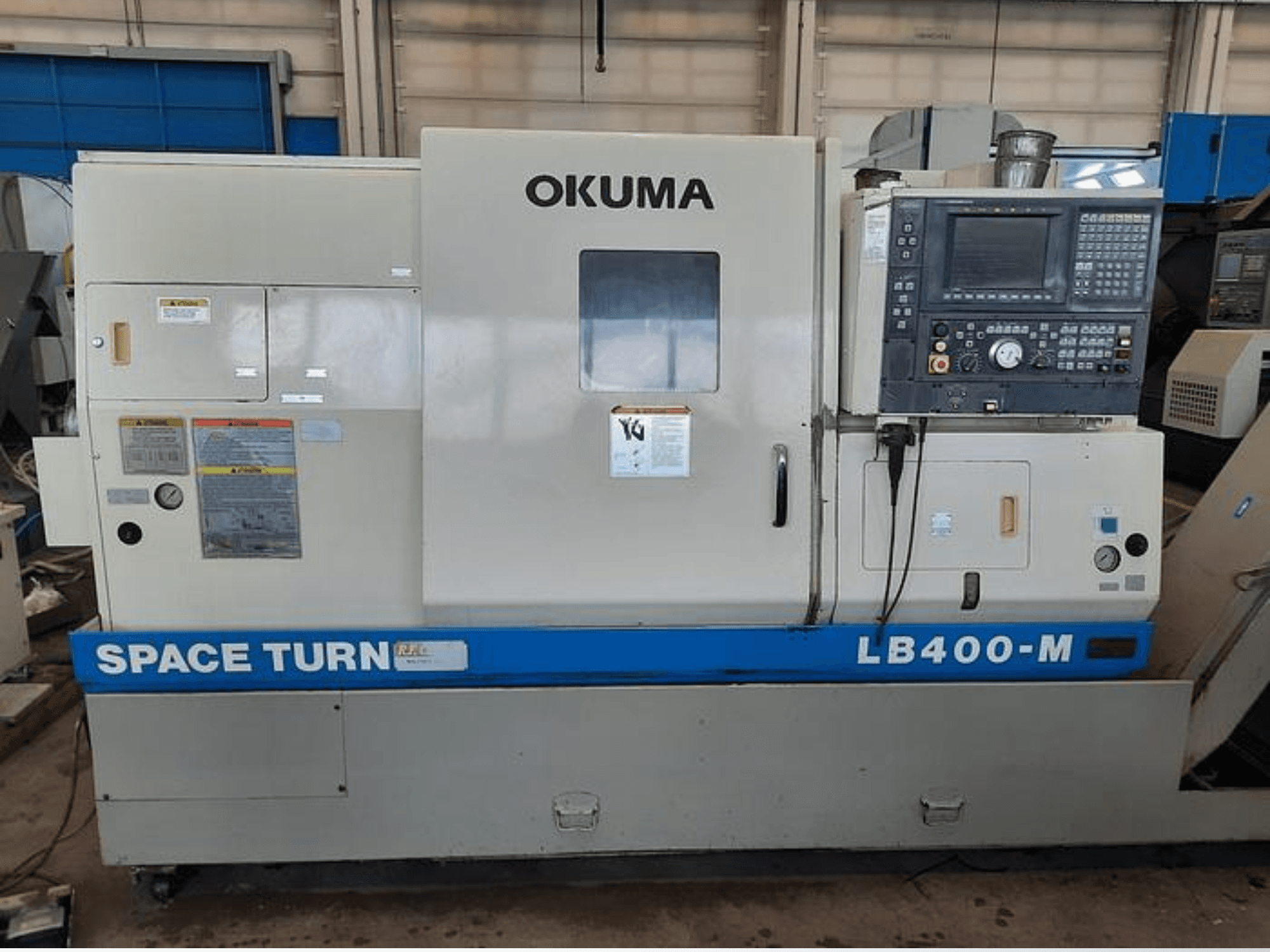 Torno CNC Okuma LB400-M, vista frontal com painel de controlo, etiquetas de aviso e a marca azul "SPACE TURN" visível.