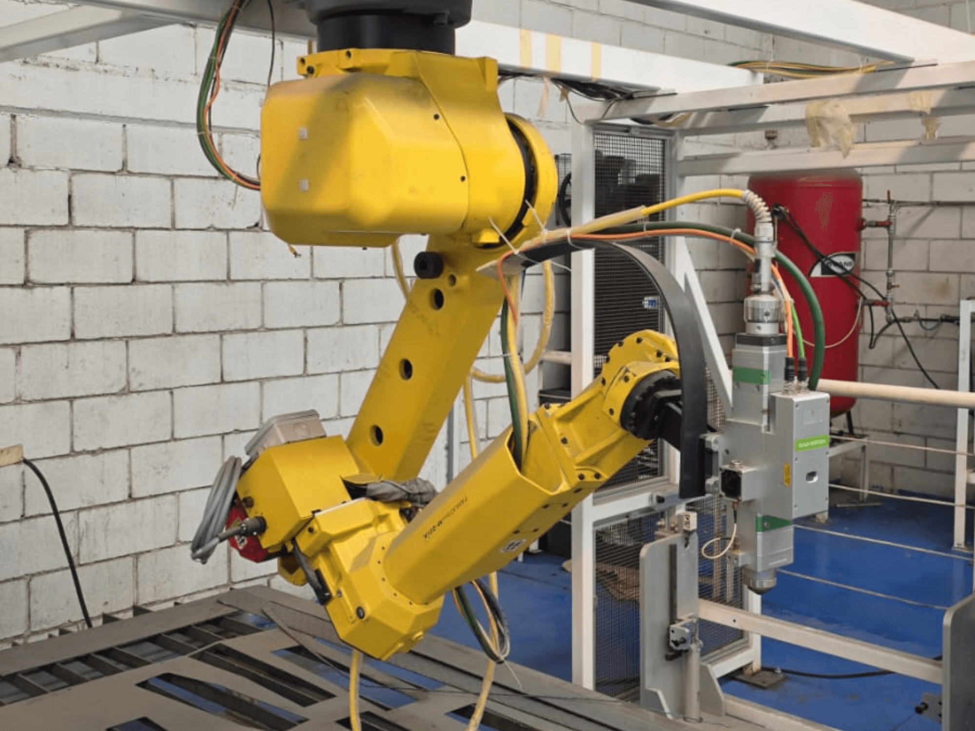 Braço robótico amarelo com uma ferramenta de precisão, com várias articulações e cabos, visto de um ângulo frontal num ambiente industrial.