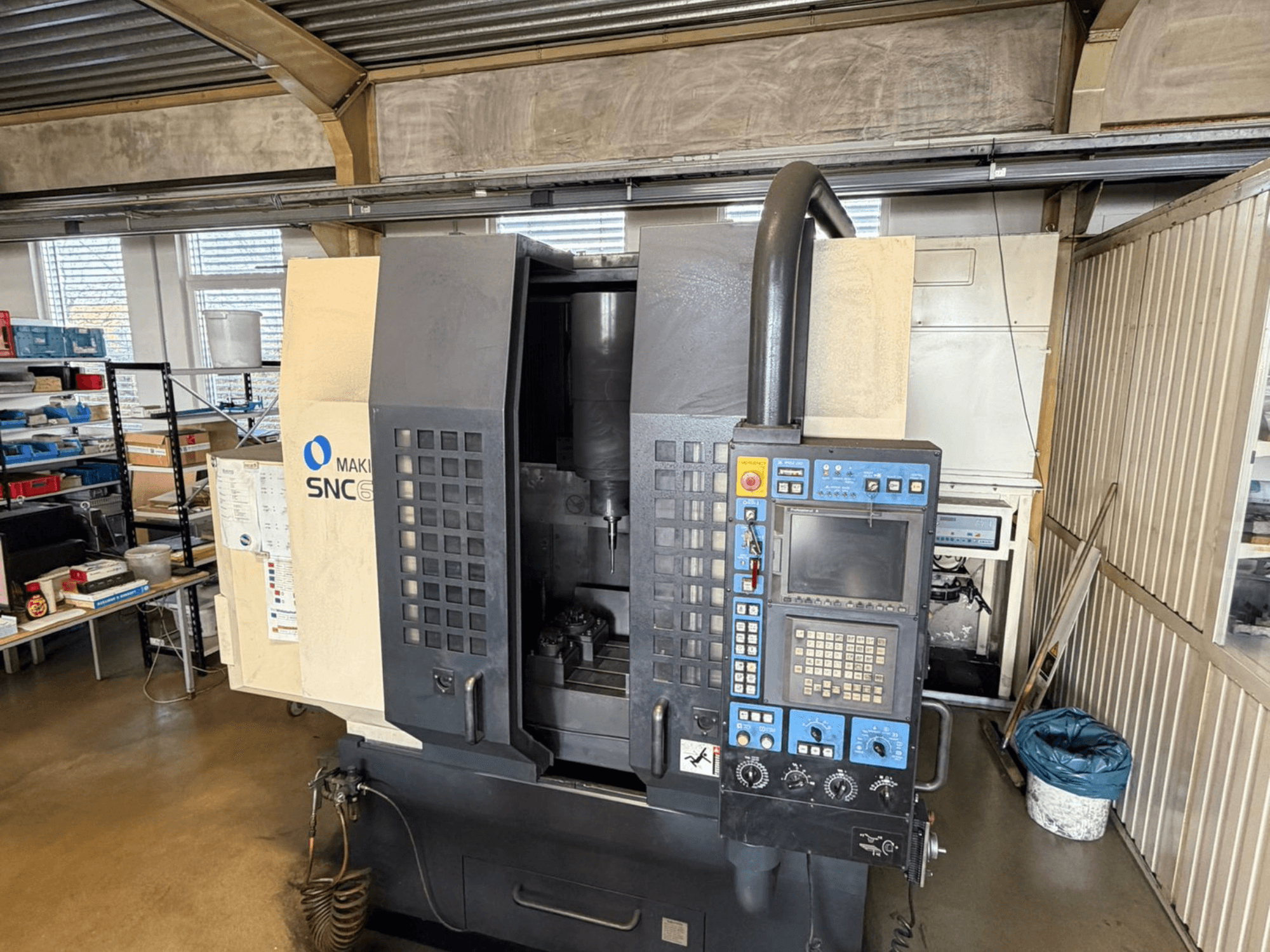 Centro de maquinação vertical Makino SNC 64-A15 em vista frontal, mostrando o painel de controlo, a área de maquinação e o espaço de trabalho industrial.