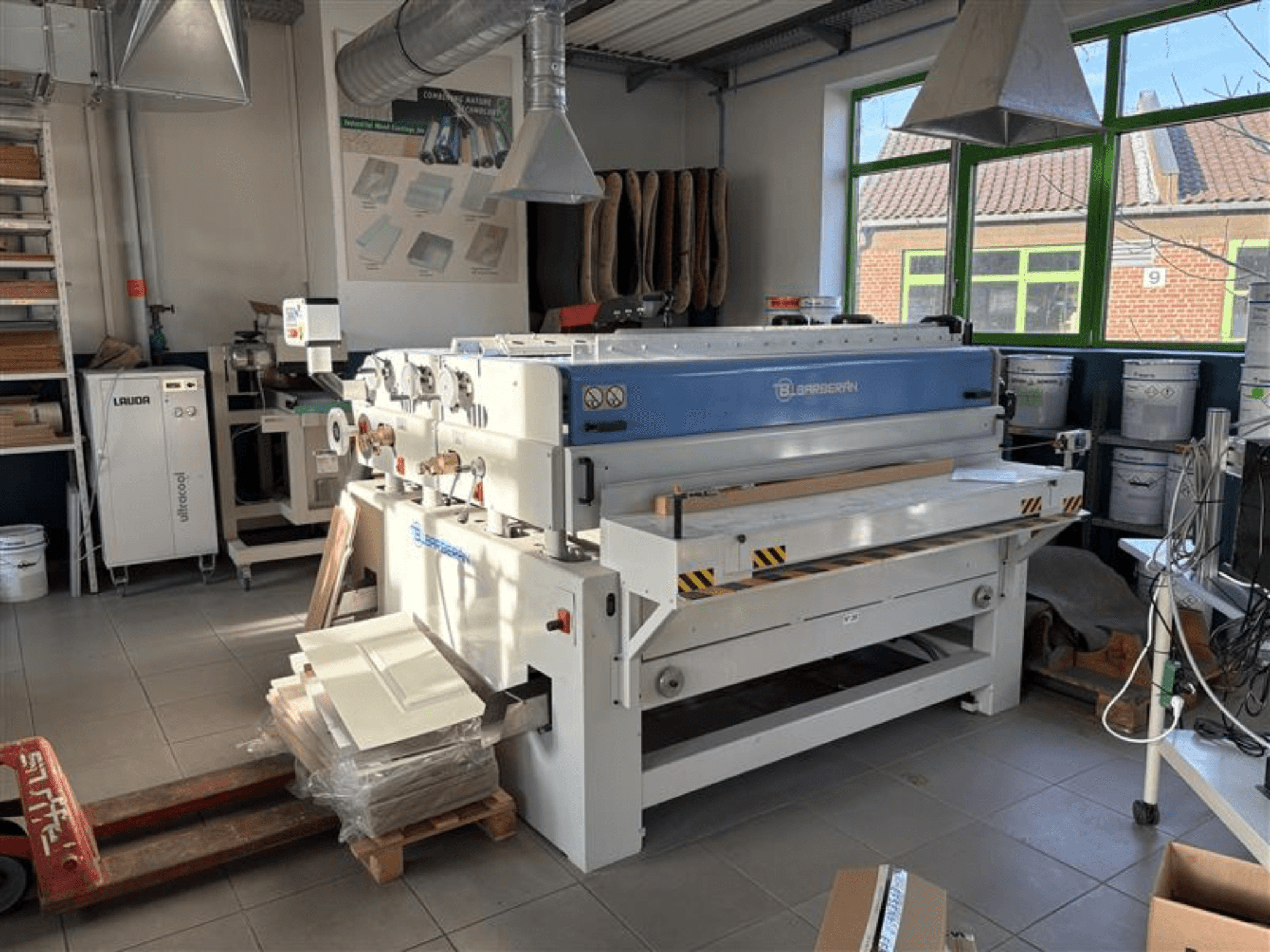 Máquina de trabalhar madeira Blaserbn numa oficina, vista frontal; com painéis de madeira numa palete e equipamento moderno em segundo plano.