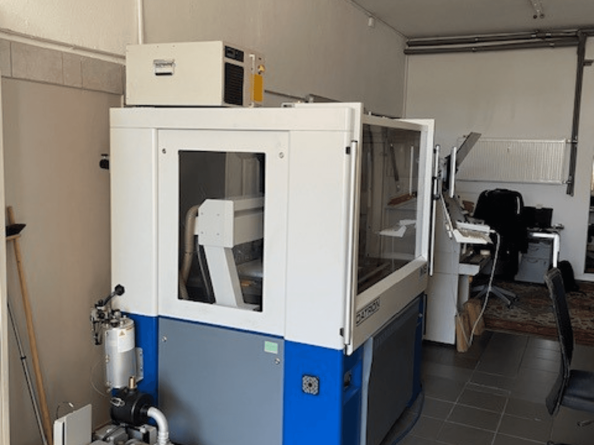 Máquina industrial azul e branca com painéis transparentes, vista frontal, inclui acesso de controlo e unidade de ventilação visível.