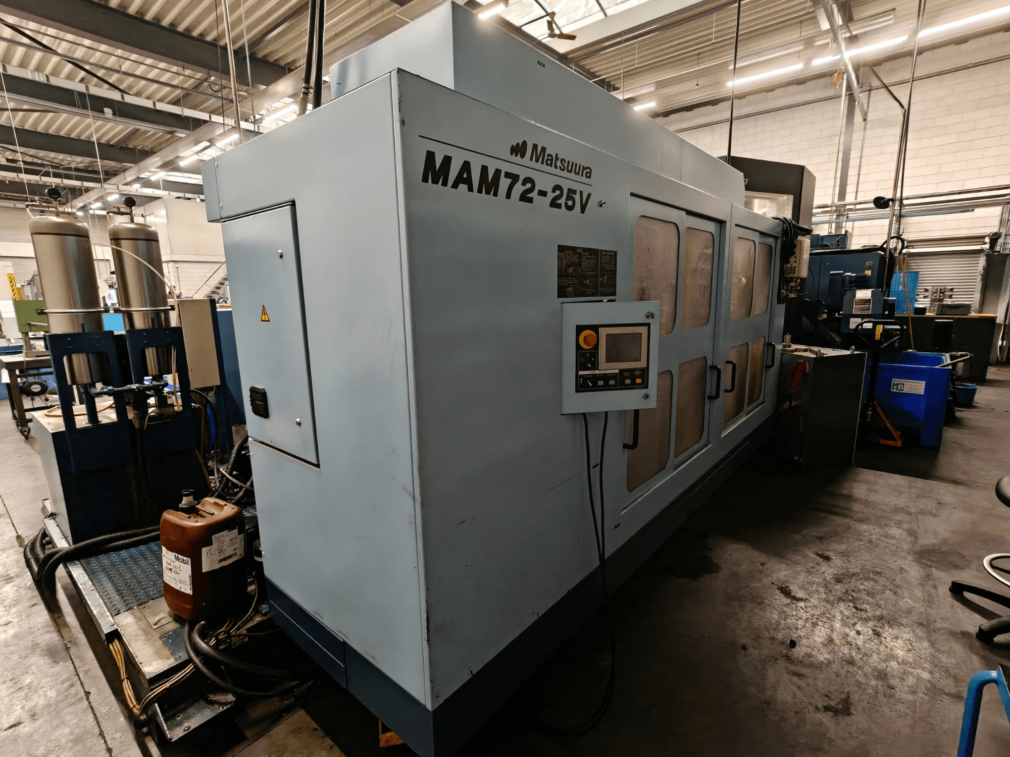 Máquina CNC Matsuura MAM72-25V, vista lateral, com painel de controlo, dois depósitos de refrigeração e espaço de trabalho num ambiente industrial.