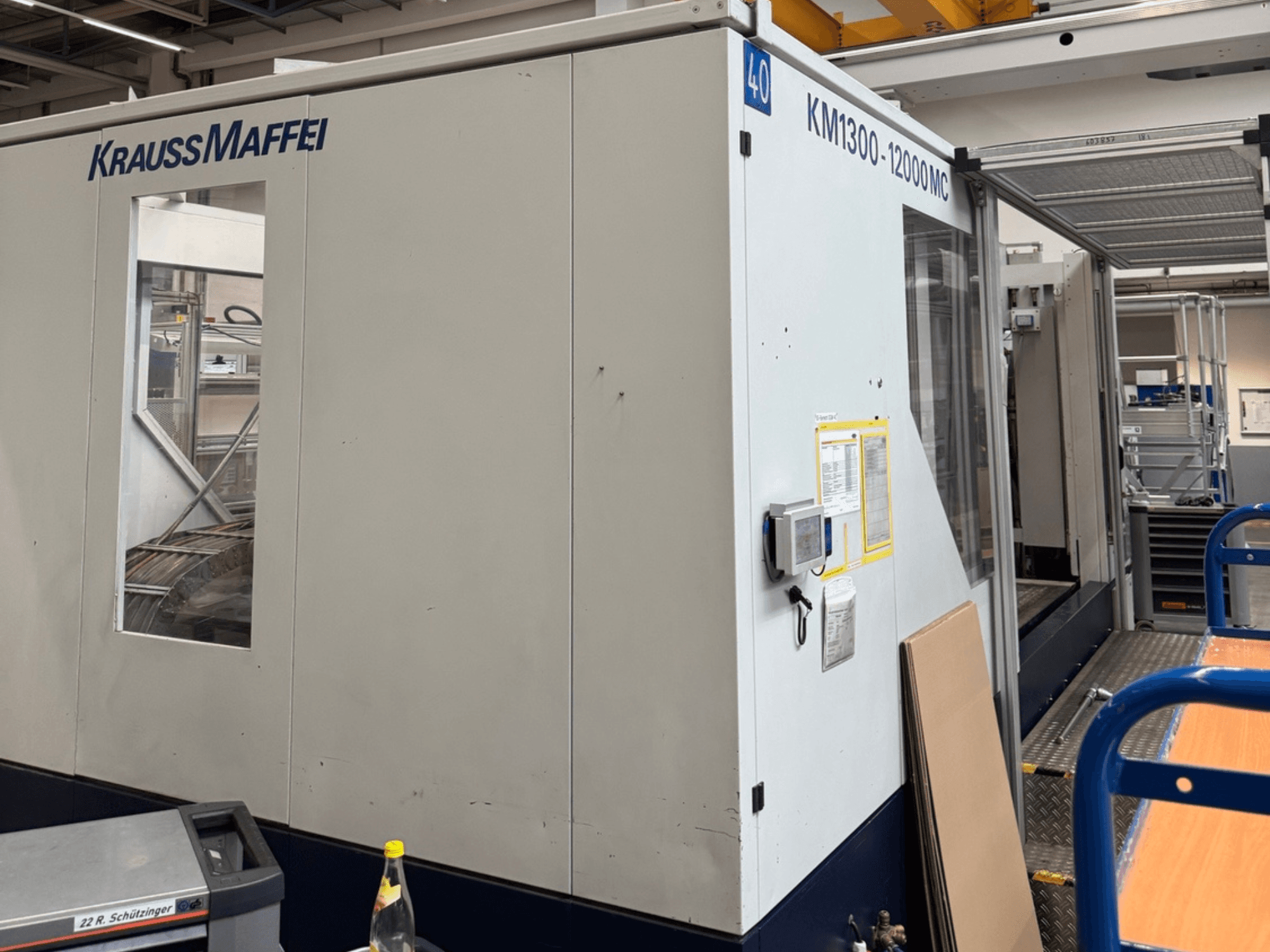 Máquina de moldagem por injeção hidráulica Krauss Maffei KM1300-12000 MC, vista lateral, mostrando o painel de controlo e o espaço operacional.