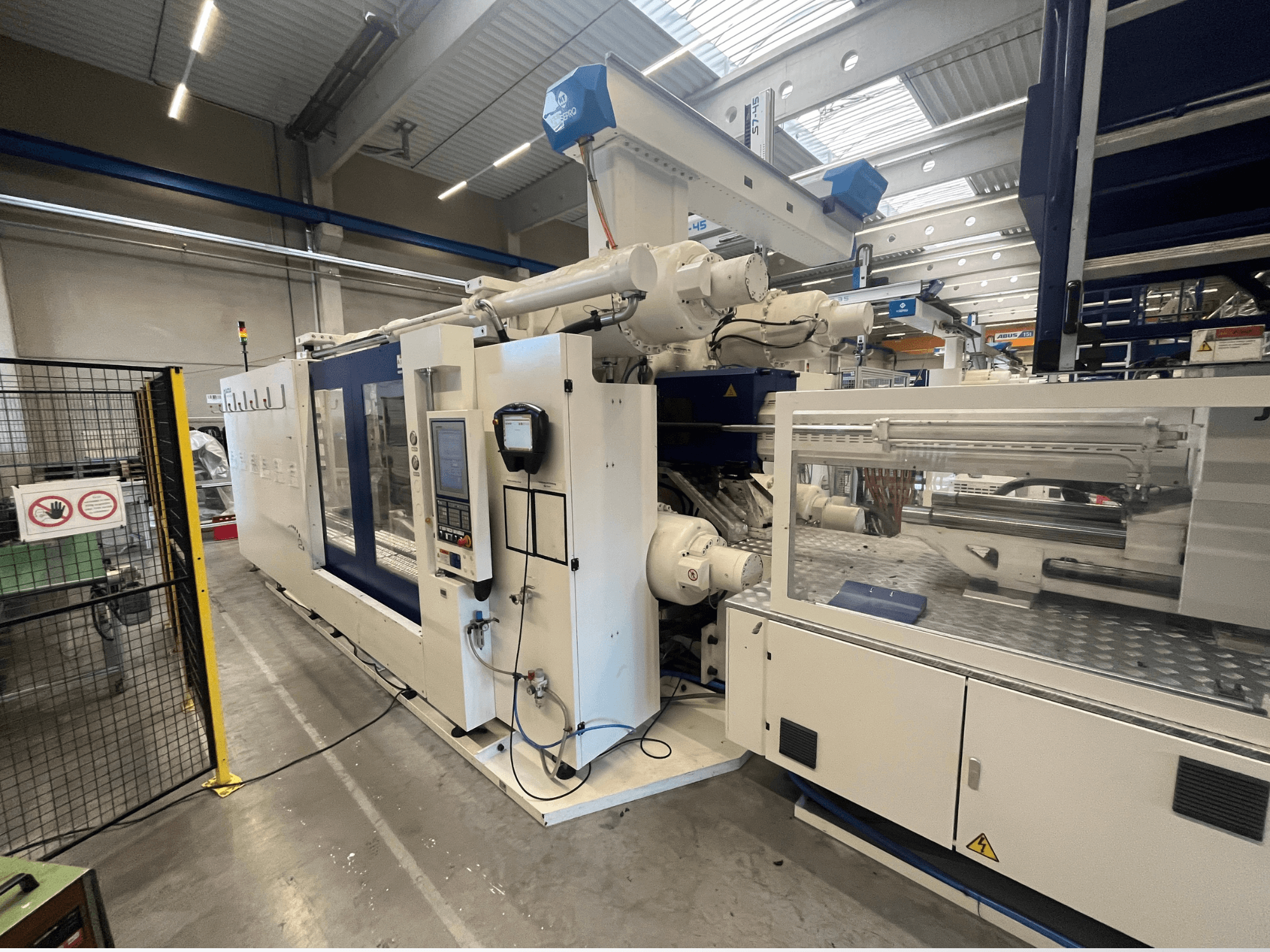 Máquina de moldagem por injeção em branco e azul, vista frontal, com painel de controlo, braço robótico e sinalização de segurança.