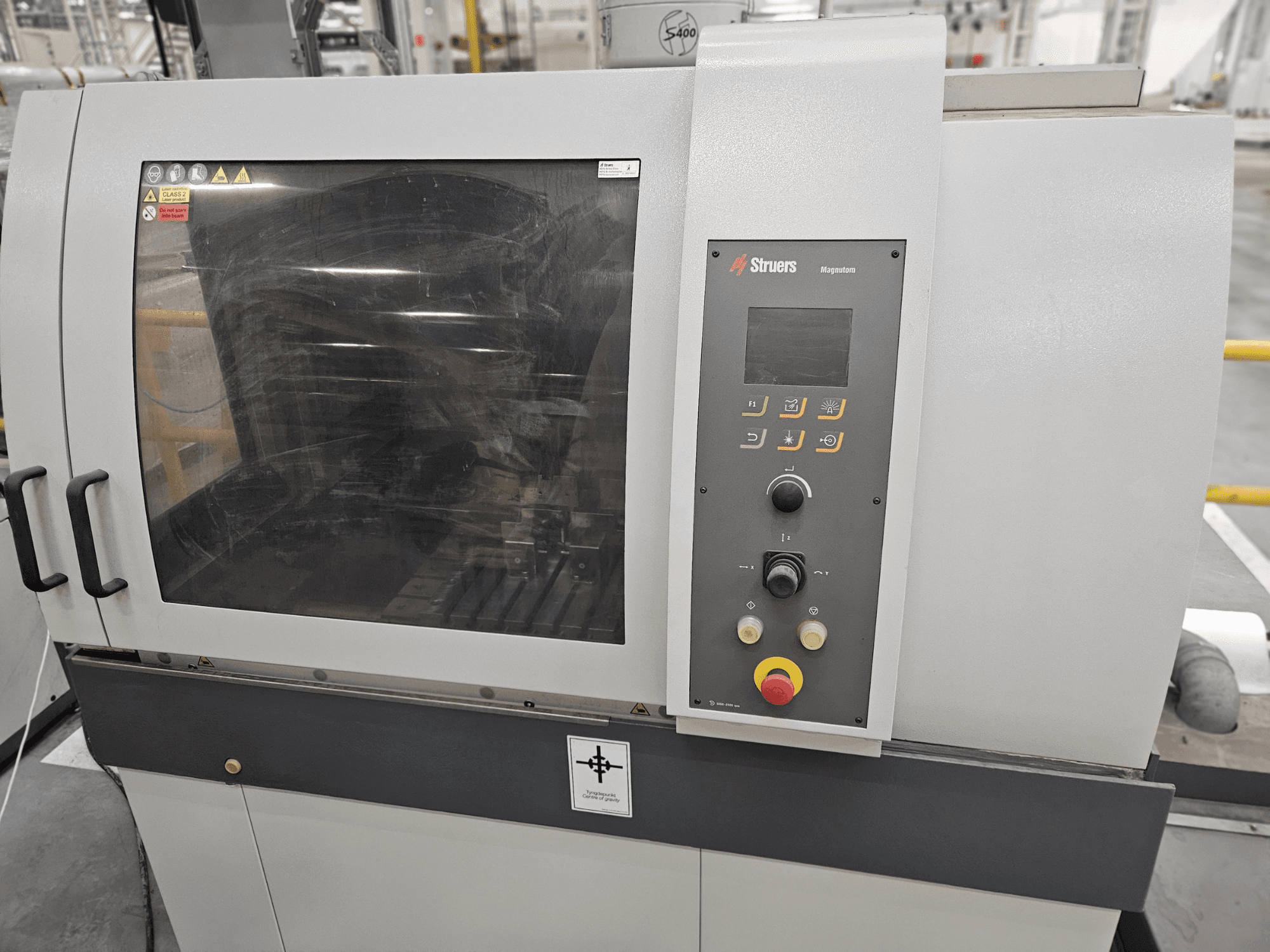 Vista frontal da máquina Struers Magpentor com painel de controlo, botões e uma grande janela de visualização. Etiquetas técnicas visíveis.