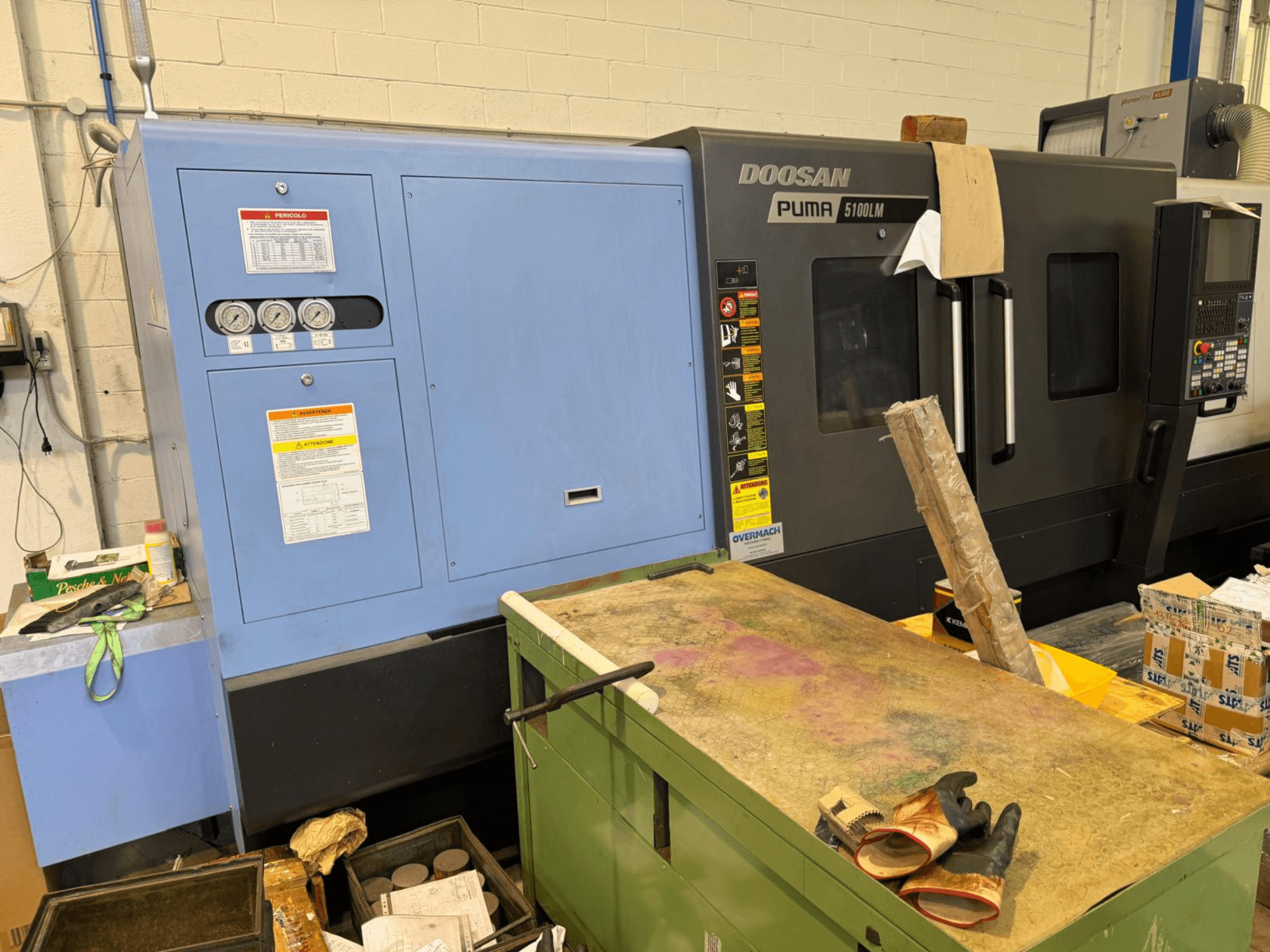 Painel de controlo e máquina CNC Doosan Puma 5100LM, vista lateral. Indicadores e etiquetas de aviso visíveis na unidade de controlo azul.