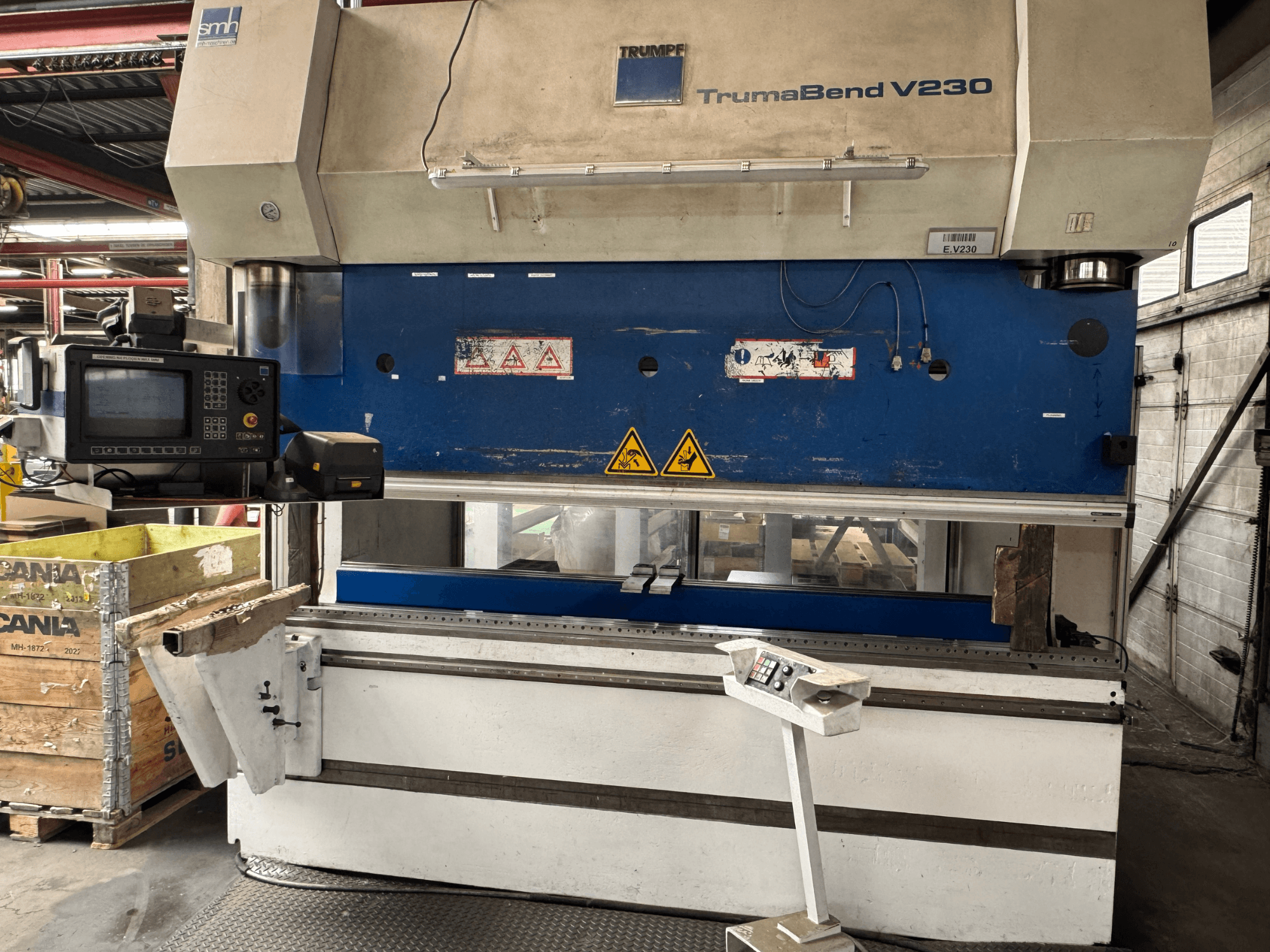 Prensa dobradeira TRUMPF TrumaBend V230, vista frontal com painel de controlo, etiquetas de aviso e um braço de suporte da peça de trabalho visíveis.