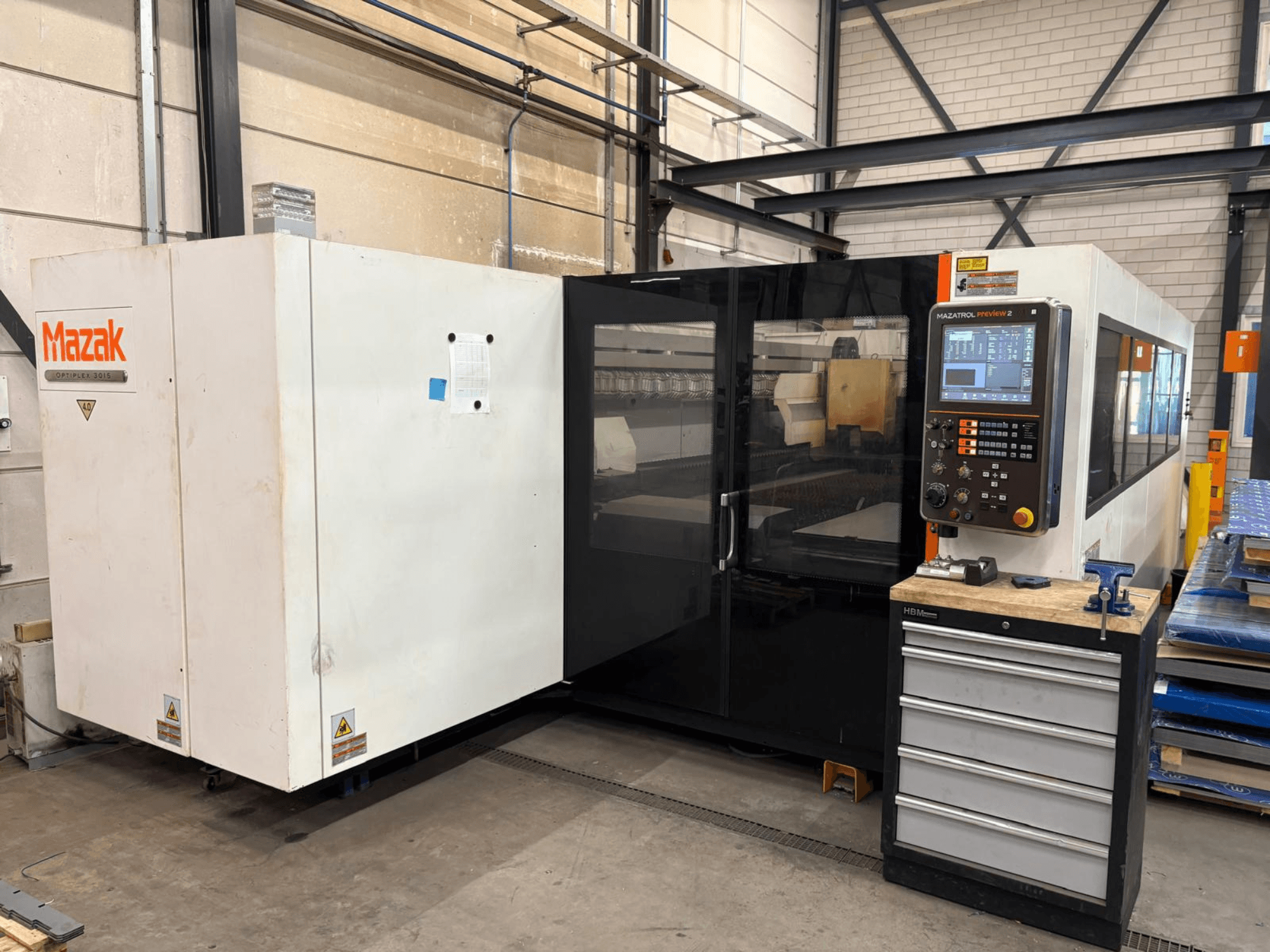 Máquina de corte a laser de CO₂ Mazak Optiplex 3015, vista frontal, mostrando o painel de controlo e o ambiente industrial.