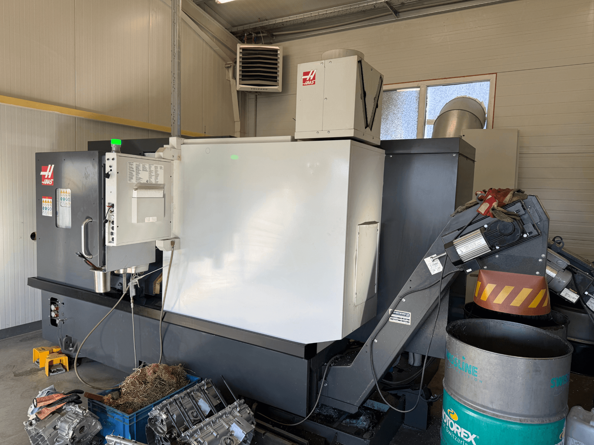 Máquina CNC Haas com painel de controlo, vista lateral; linhas de refrigeração e de óleo visíveis; restos de metal no contentor azul; ambiente industrial.