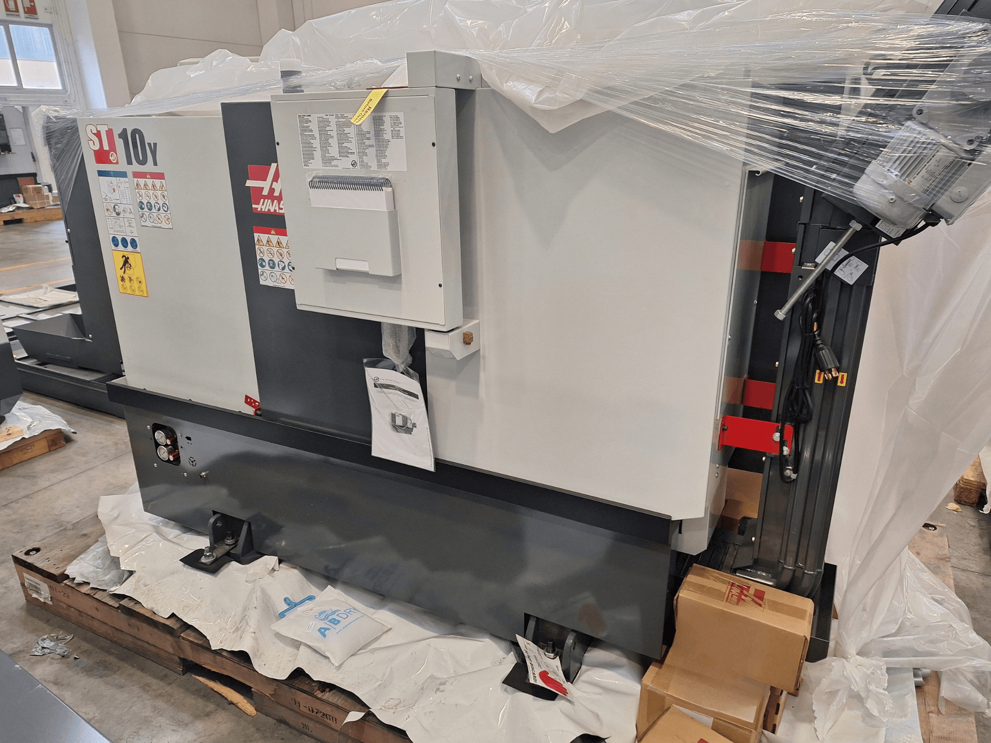 Vista lateral da máquina de torneamento horizontal HAAS ST-10Y EU, embalada em plástico, com o painel de controlo e as etiquetas de aviso visíveis.