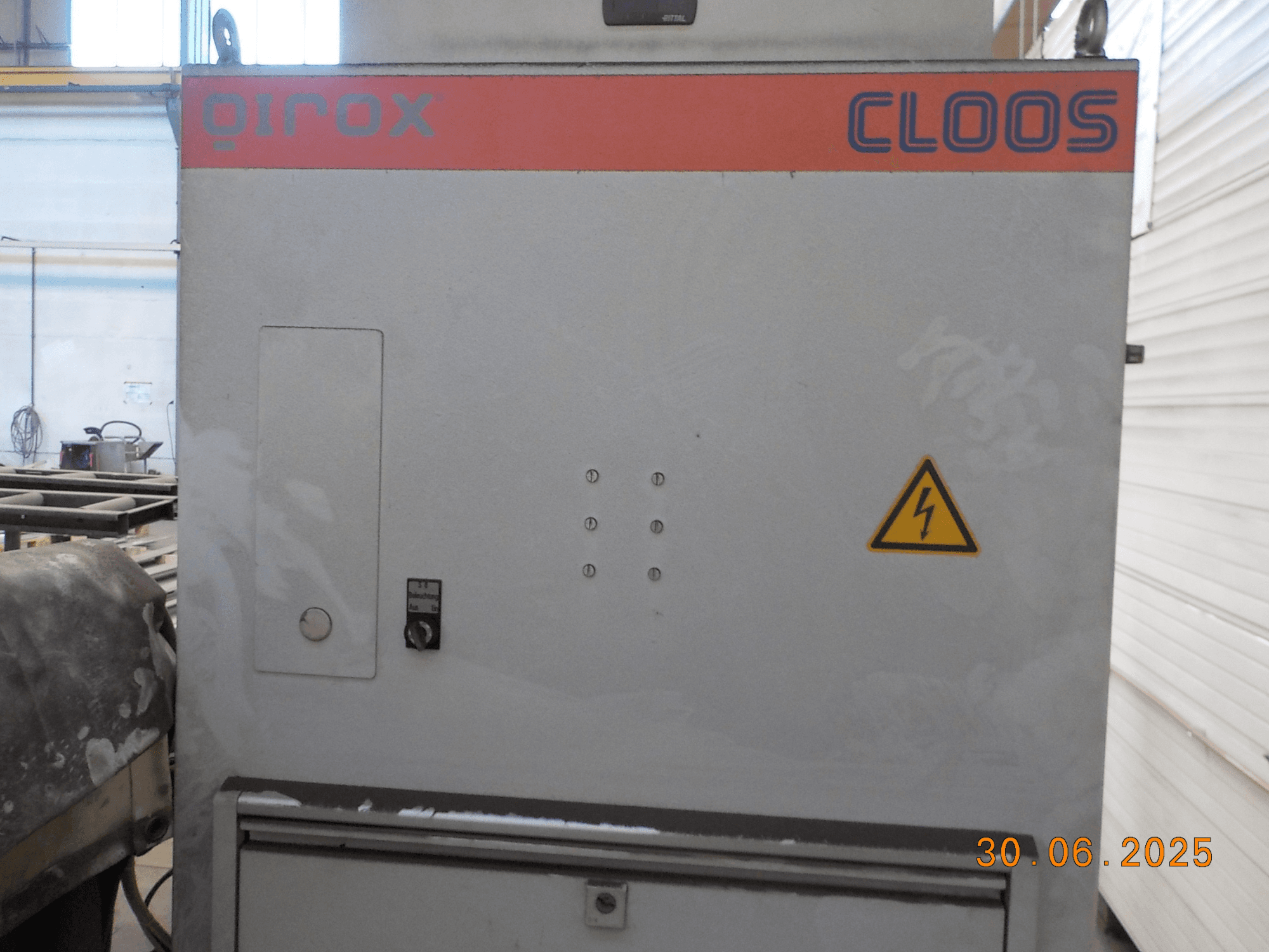 Vista lateral da máquina de soldar Cloos QIROX, com painel de controlo e sinal de aviso, com uma superfície empoeirada e carimbo de data visível.