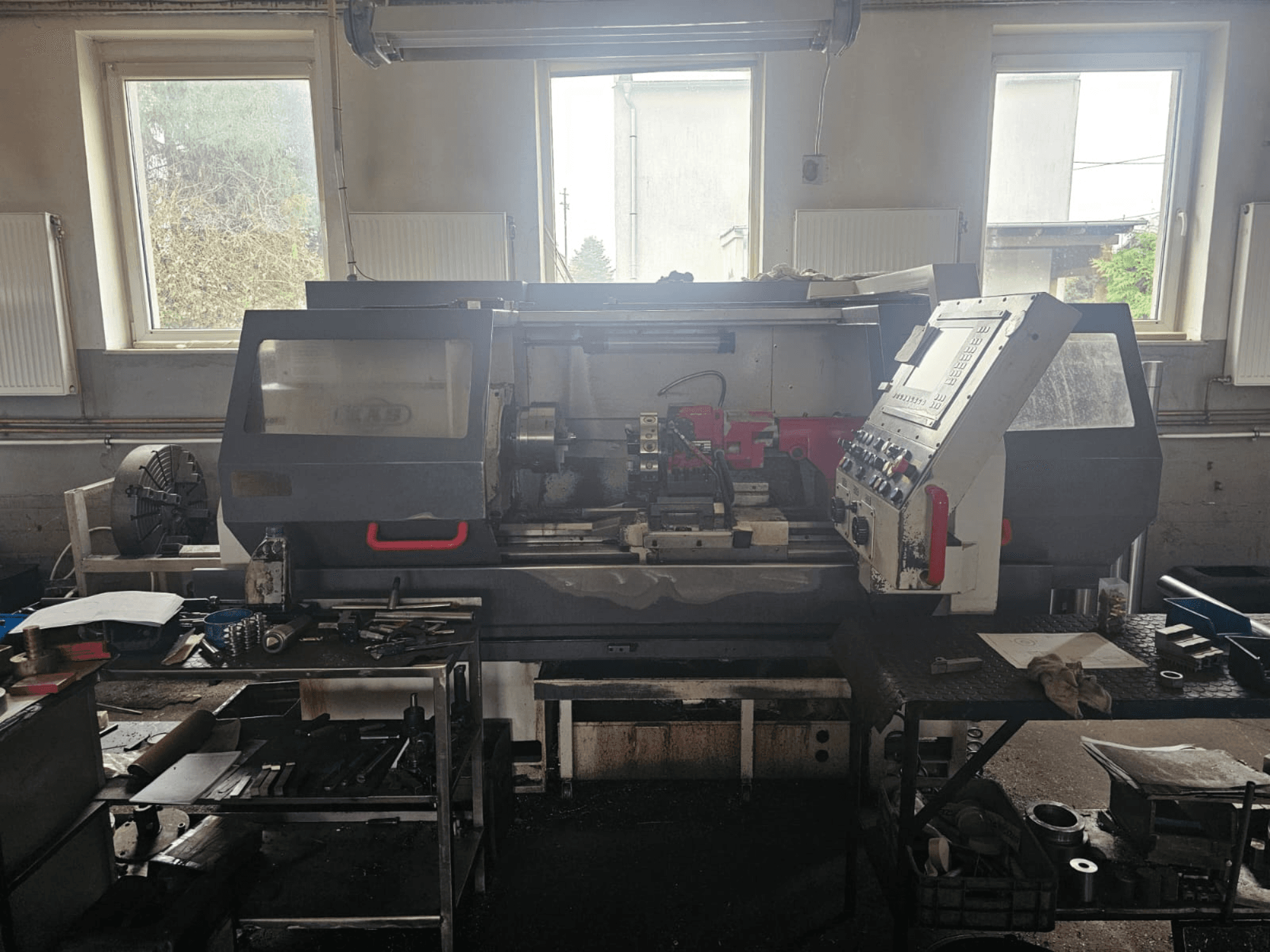 Torno mecânico numa oficina, vista frontal, com painel de controlo e ferramentas em cima de mesas, rodeado por janelas e um espaço de trabalho desordenado.