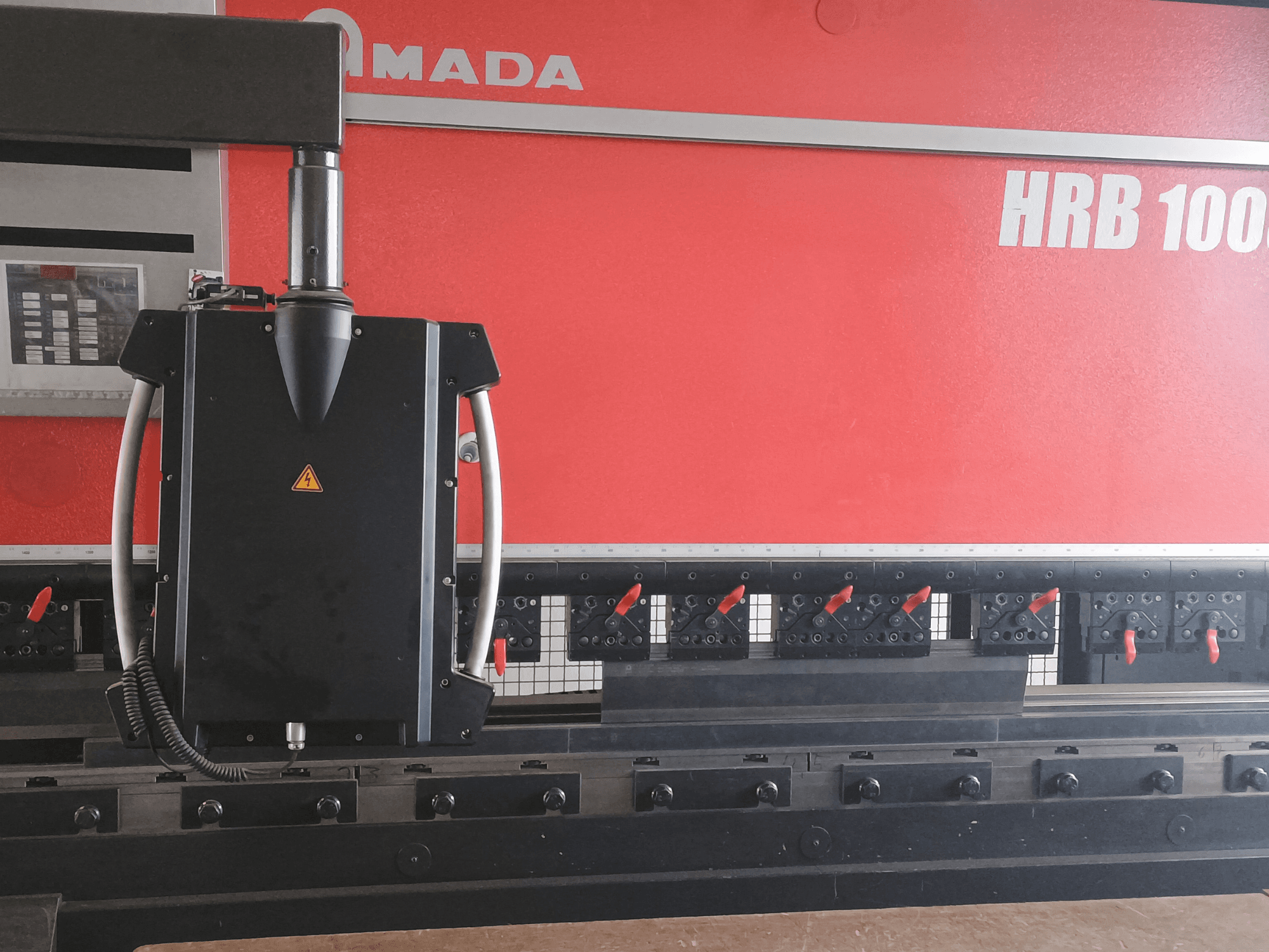 Vista frontal da máquina de dobragem Amada HRB 100; possui uma cabeça de controlo preta com um bocal cónico e pinças ajustáveis.