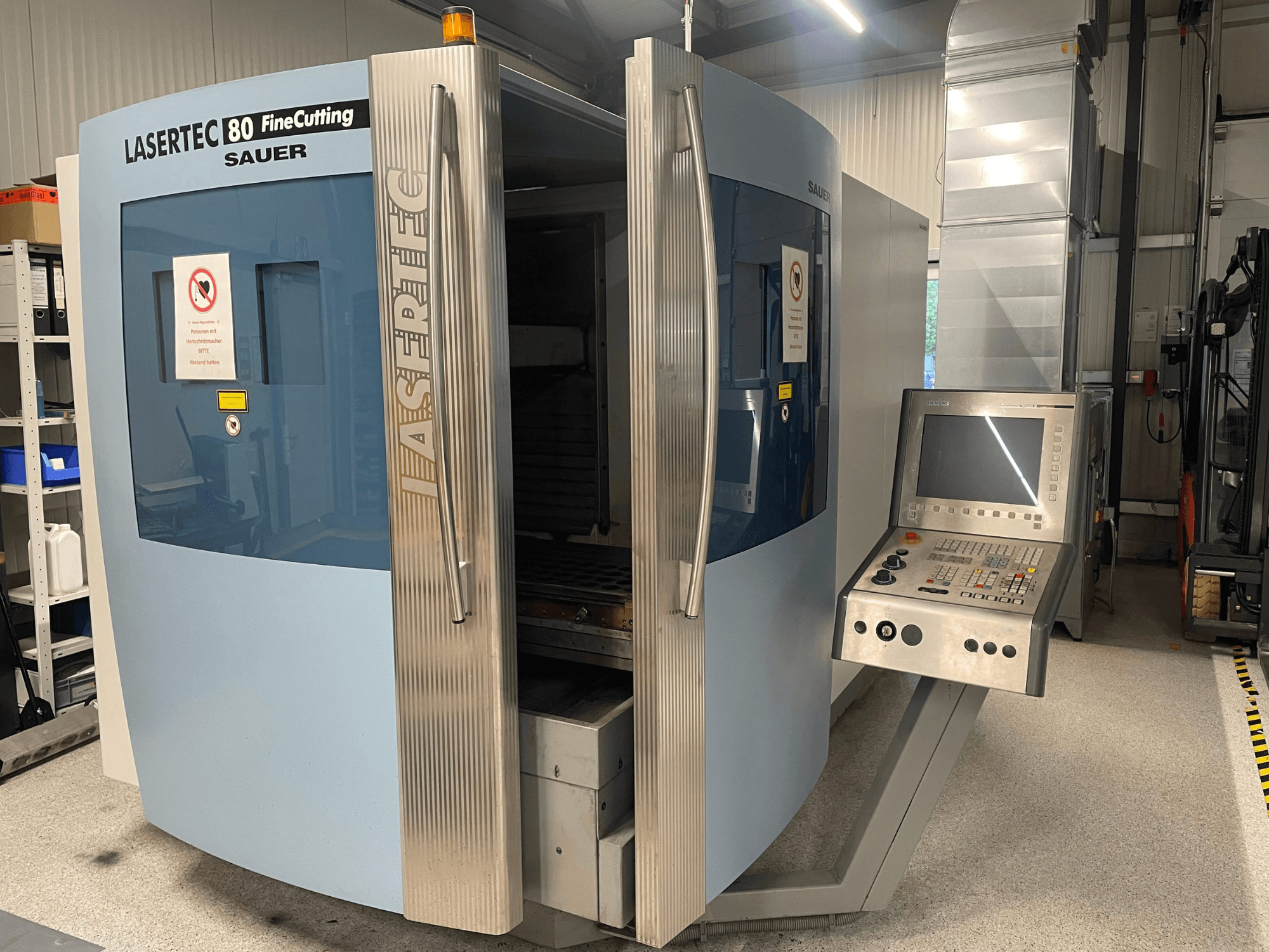Vista frontal da máquina de corte de chapa DMG Sauer Lasertec80 FineCutting, com o painel de controlo, as portas abertas e a sinalização de segurança visível.