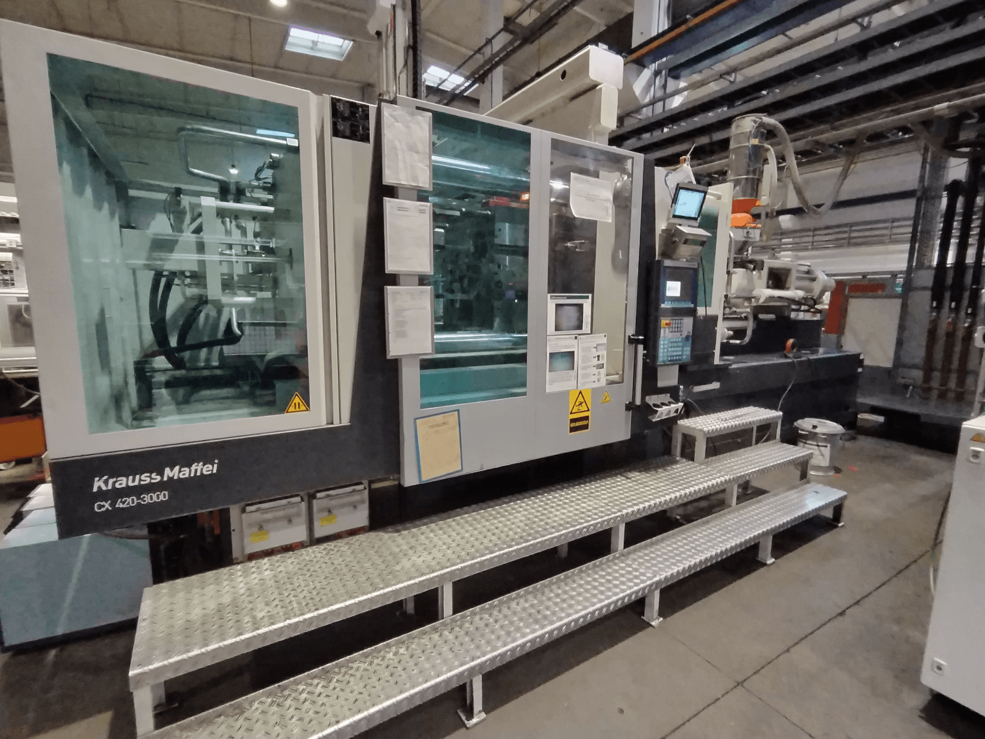 Máquina de moldagem por injeção Krauss Maffei CX 420-3000, vista lateral, com painel de controlo e sinalização de segurança visíveis, colocada em degraus metálicos.