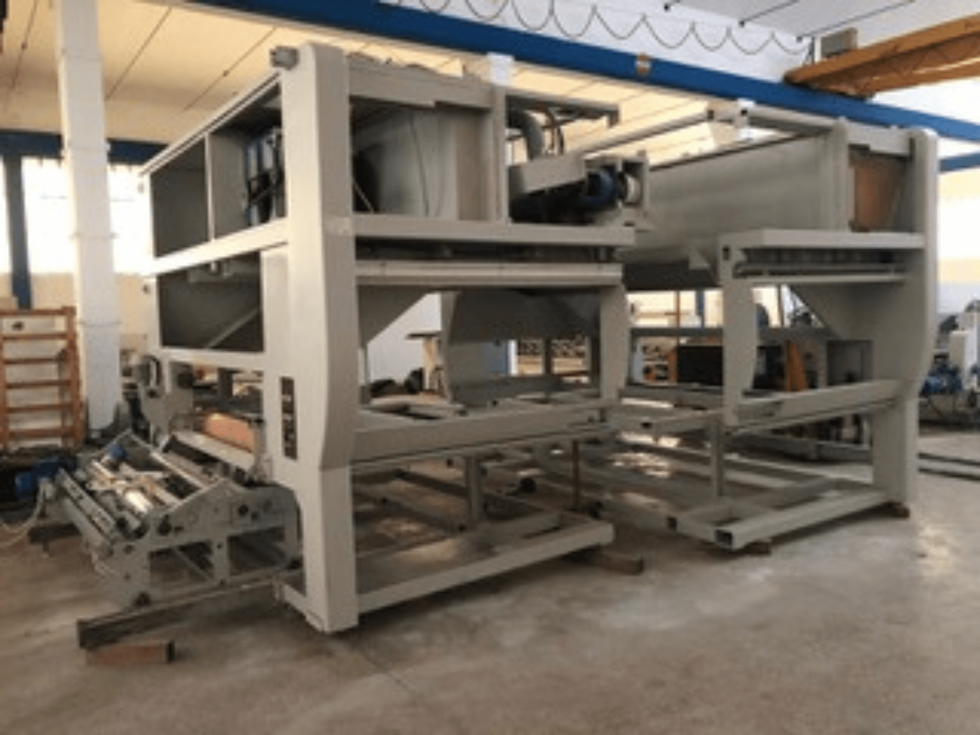 Máquina industrial com vários rolos transportadores e estrutura de grandes dimensões; vista lateral, cor cinzenta, situada numa fábrica bem iluminada.