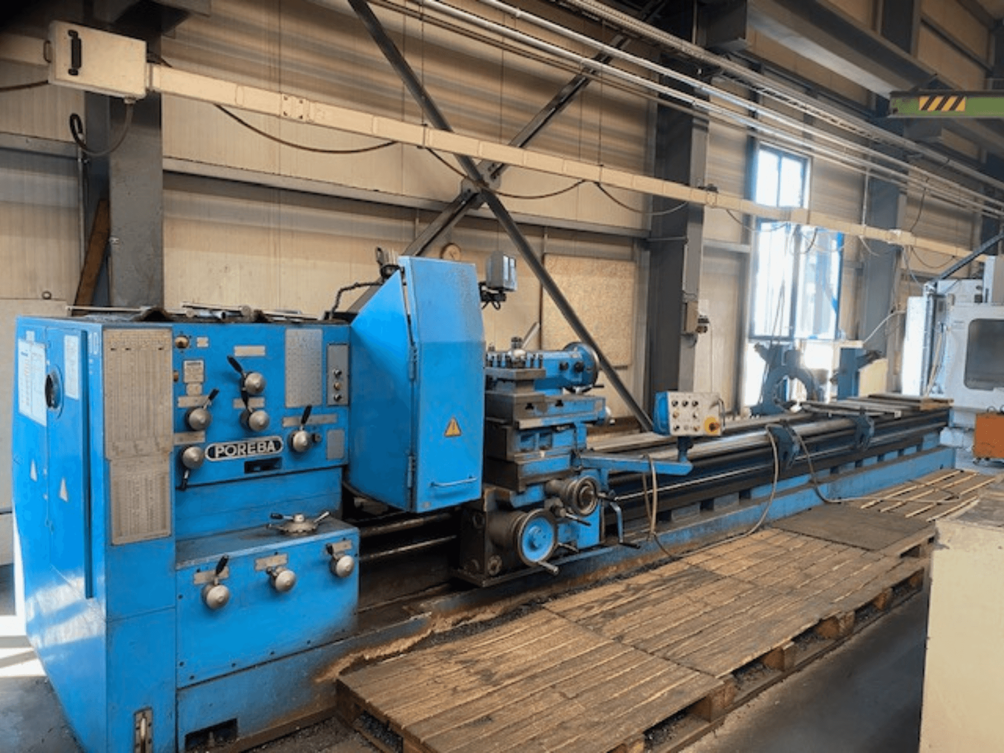 Vista frontal de uma máquina de torno POREBA azul, com botões de controlo, um descanso de ferramenta e paletes de madeira por baixo.