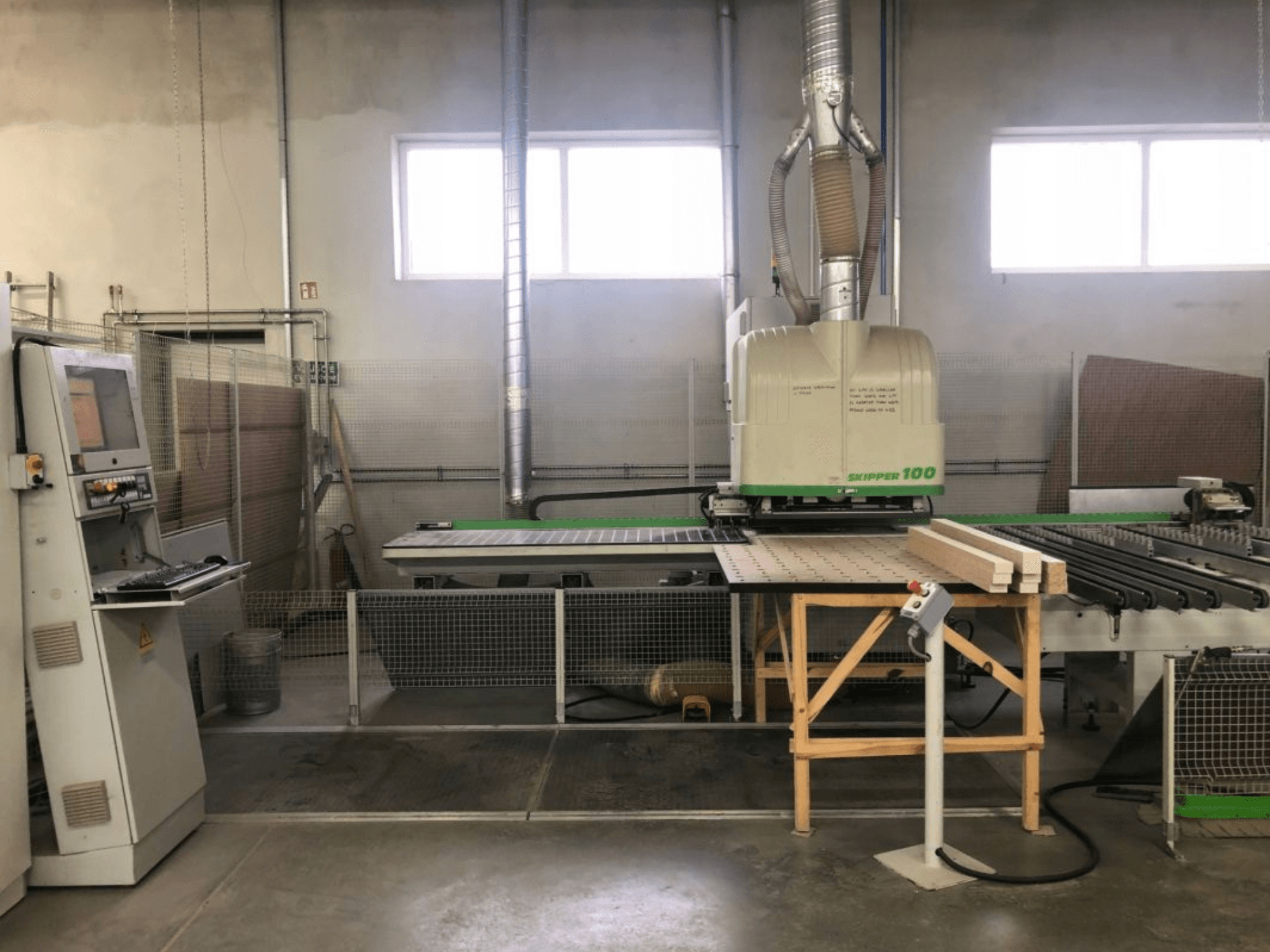 Centro de usinagem CNC BIESSE SKIPPER 100 numa oficina; a vista frontal mostra a máquina com o painel de controlo e a madeira na mesa.
