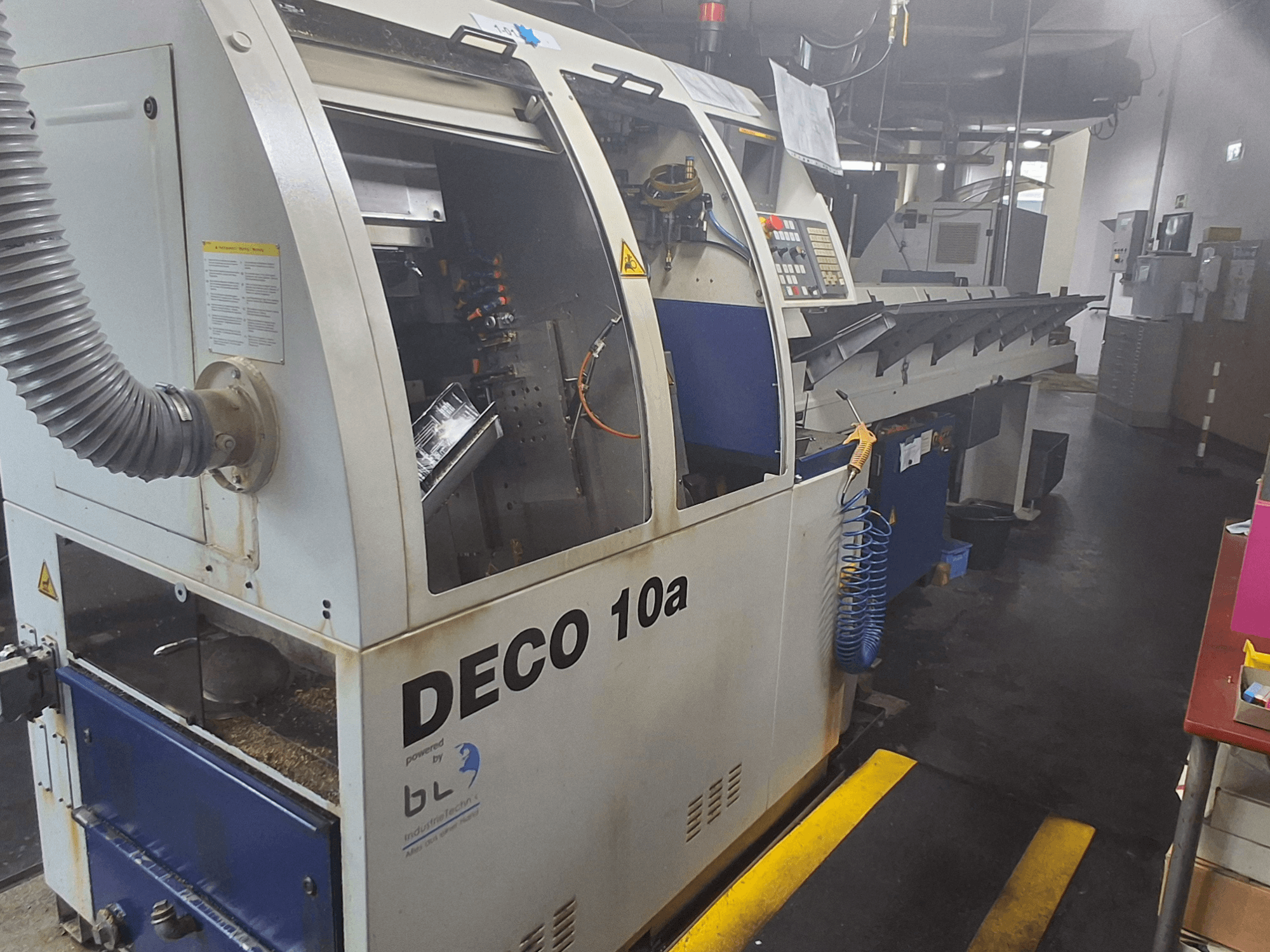 Torneiro de tipo suíço TORNOS DECO 10a, vista frontal; possui um painel de controlo, mecanismos visíveis e um tubo de extração de poeiras.
