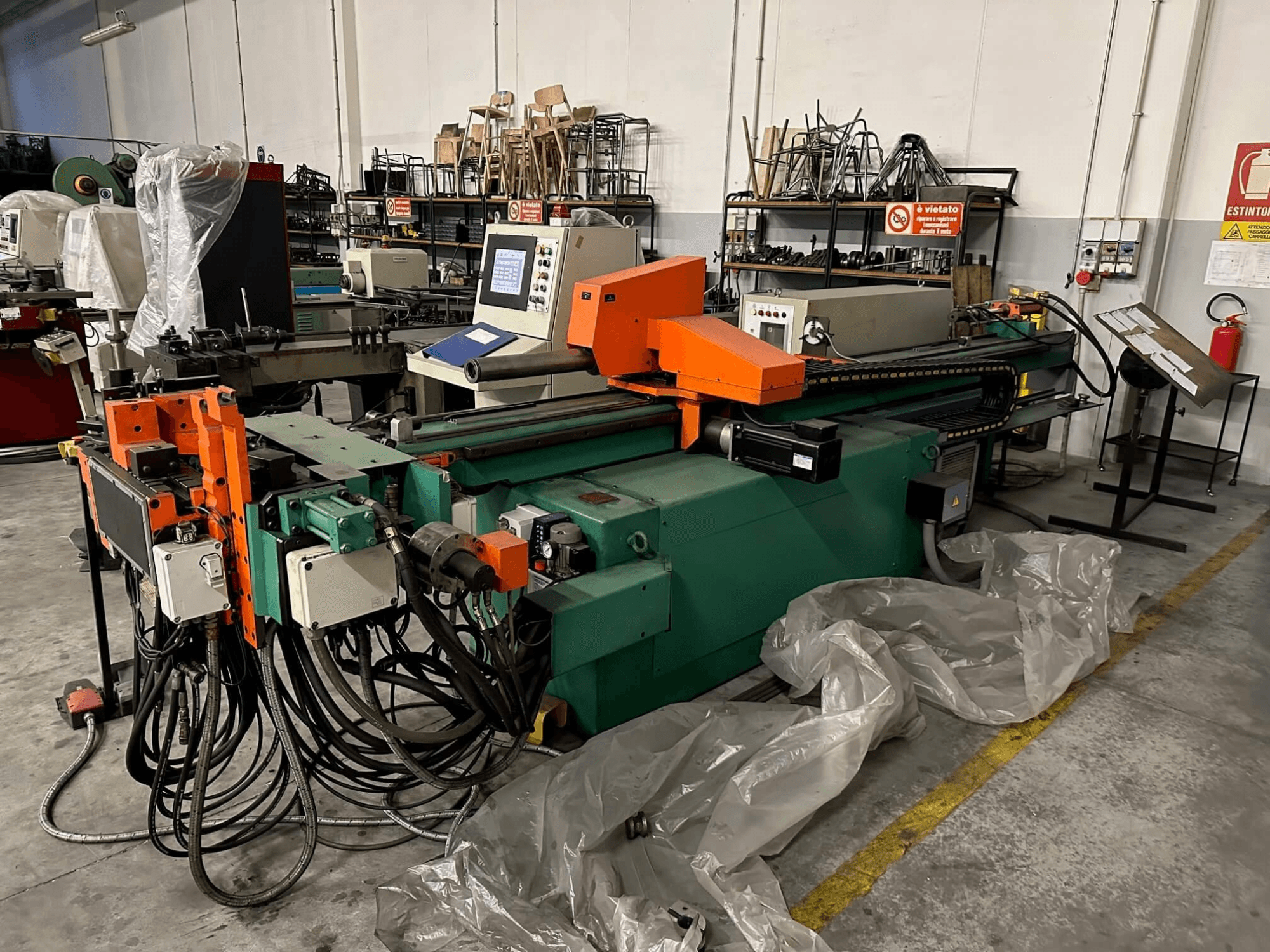 Máquina industrial com corpo laranja e verde, painel de controlo e numerosos cabos, vista de frente numa oficina.