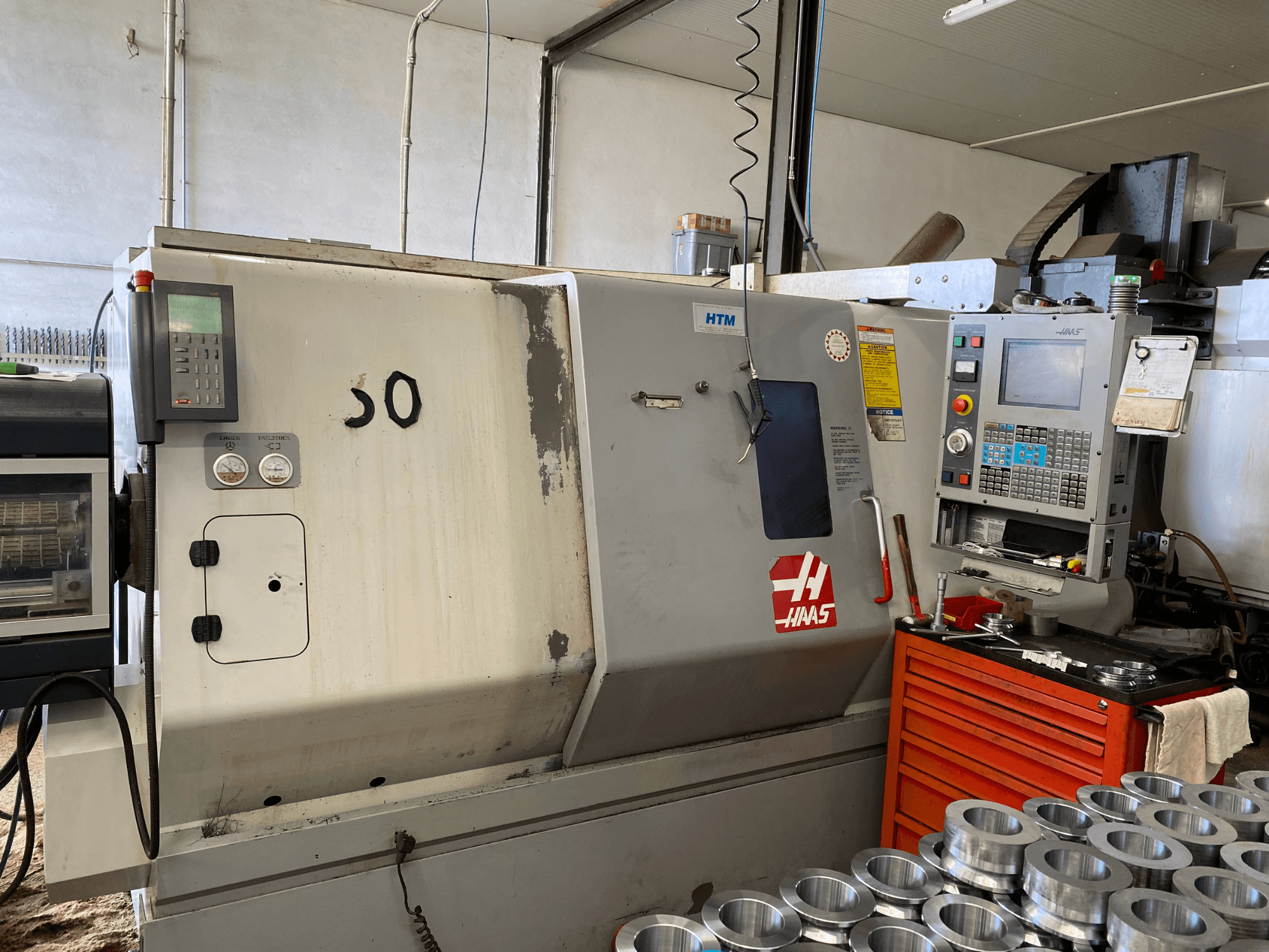 Máquina CNC Haas com painel de controlo, vista frontal, mostrando botões de operação, manómetros e peças metálicas empilhadas nas proximidades.