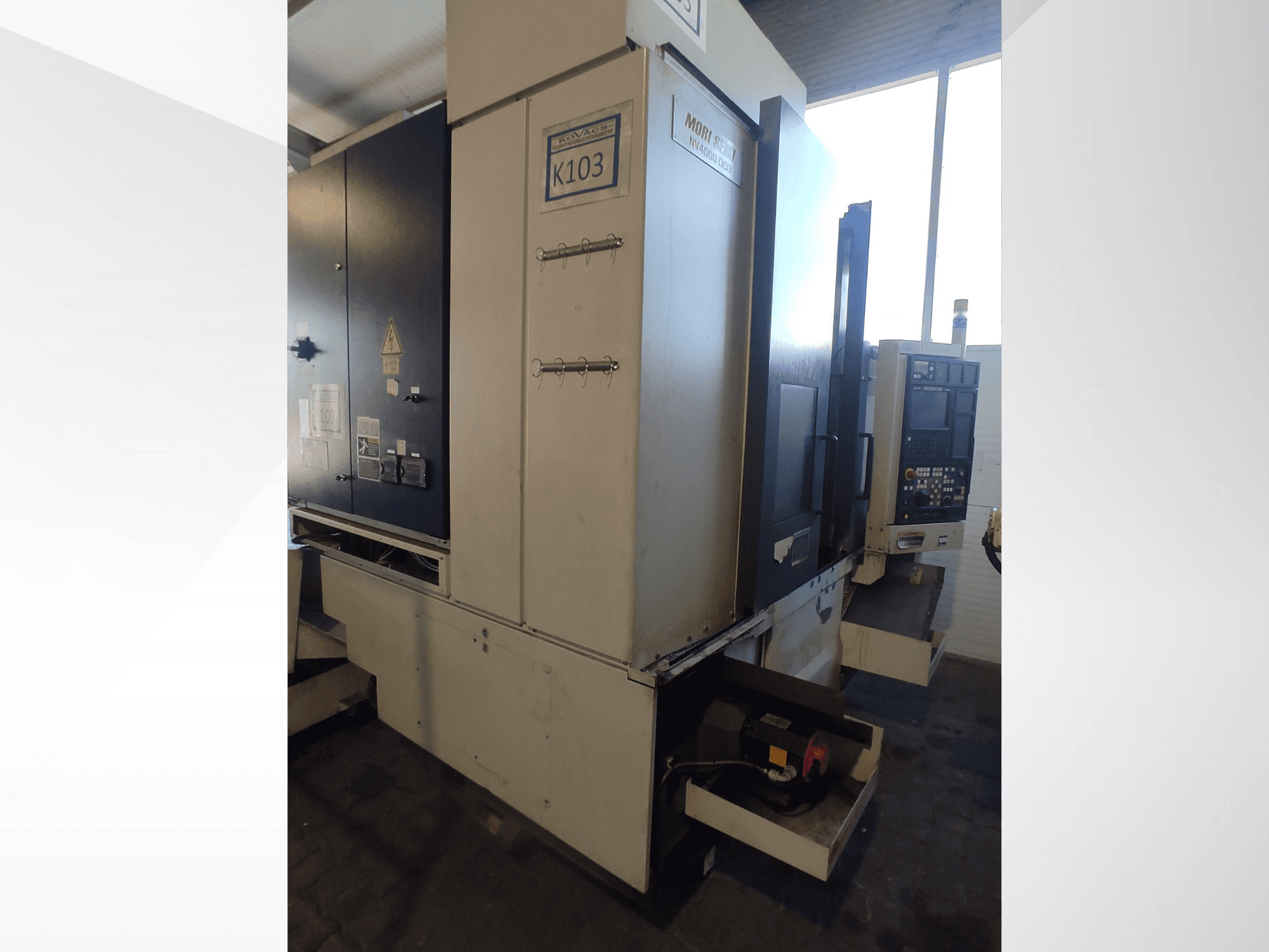 Máquina CNC Mori Seiki K103 em vista frontal, com painel de controlo, etiquetas de segurança e componentes eléctricos visíveis.