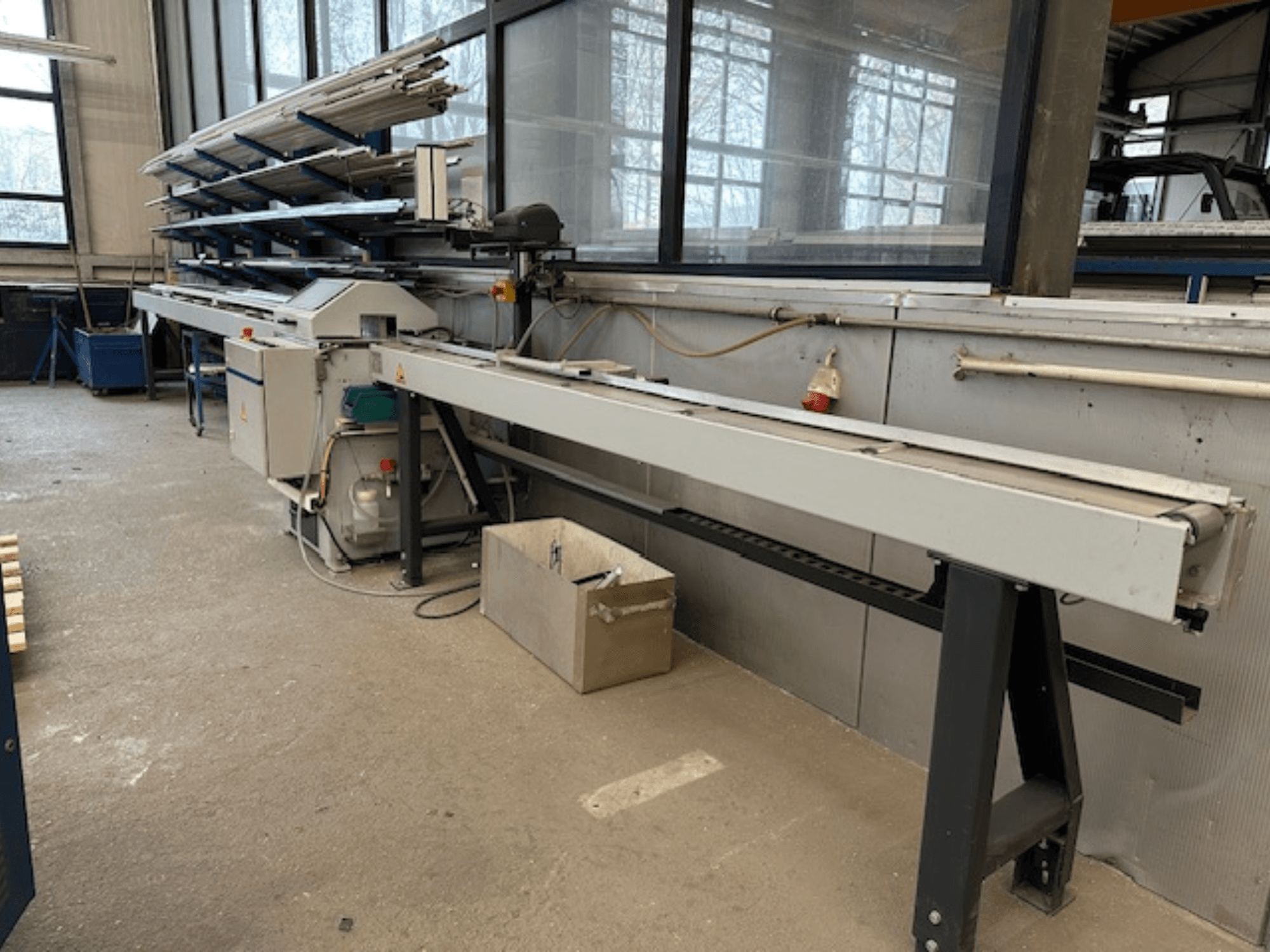 Serra circular para metal SCHÜCO PGS numa oficina, vista lateral com uma base de corte ajustável e uma prateleira de perfis metálicos por cima.