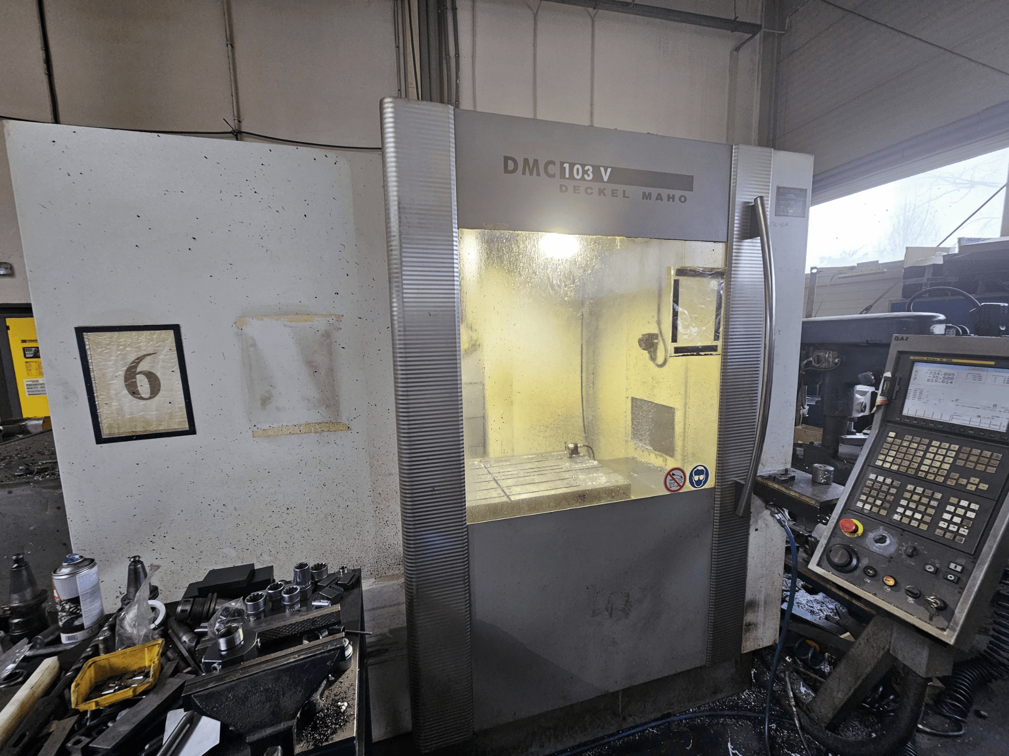 Máquina CNC Deckel Maho DMC 103 V, vista frontal, com painel de controlo e espaço de trabalho, com um número "6" na parede.