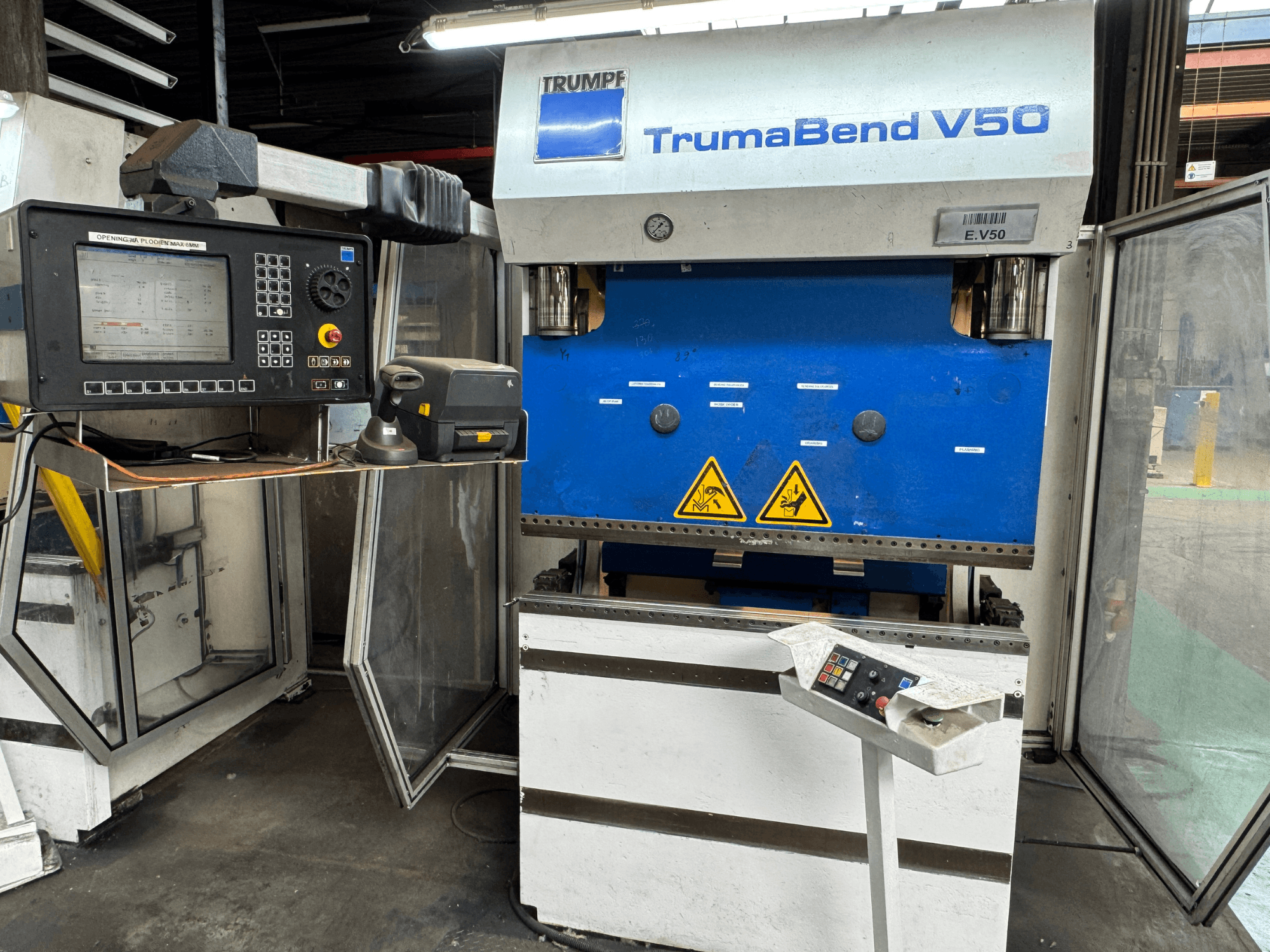 A prensa dobradeira TrumaBend V50 da TRUMPF, vista de frente, apresenta um painel de controlo, avisos de segurança e um design distinto em azul e branco.