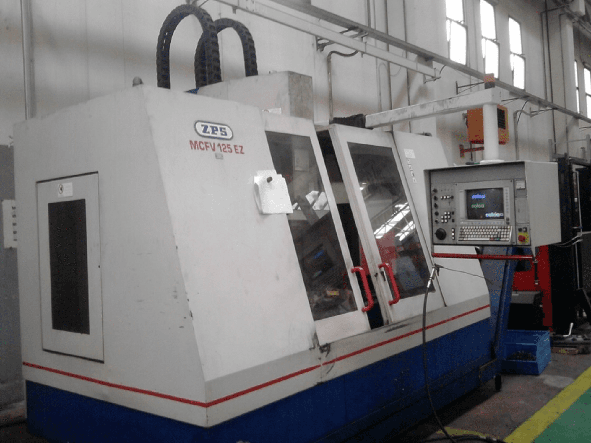 Máquina CNC ZPS MCFV 125 EZ, vista frontal com painel de controlo e punhos vermelhos, numa oficina com mangueiras visíveis.